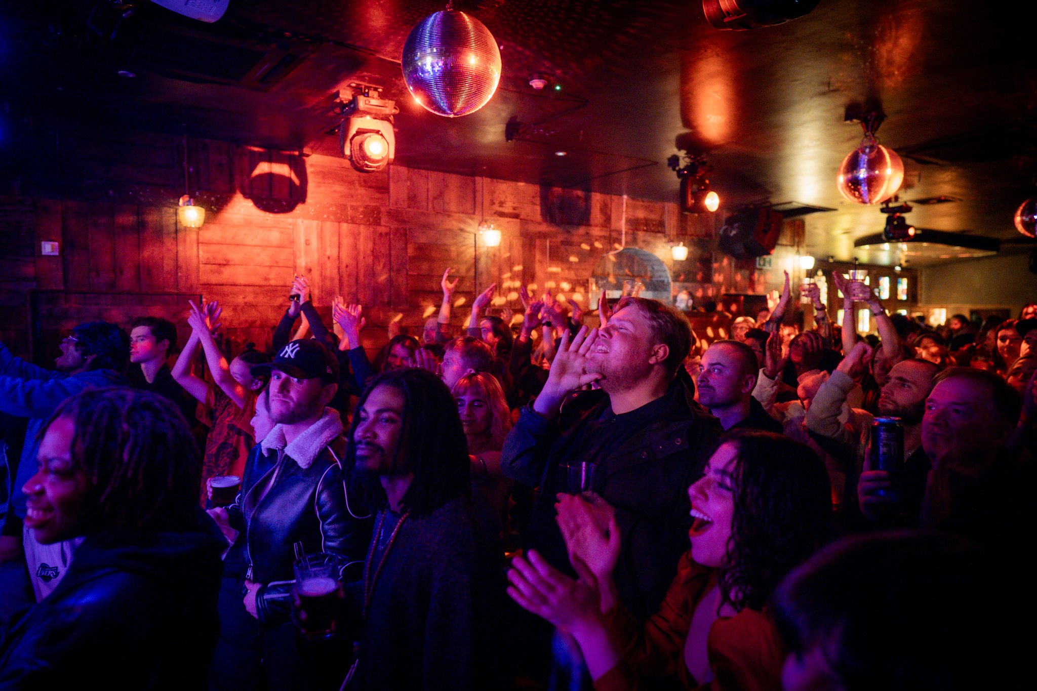 Crowd of people at a What Does Not live music night with wooden walls, disco ball, and colorful lighting.