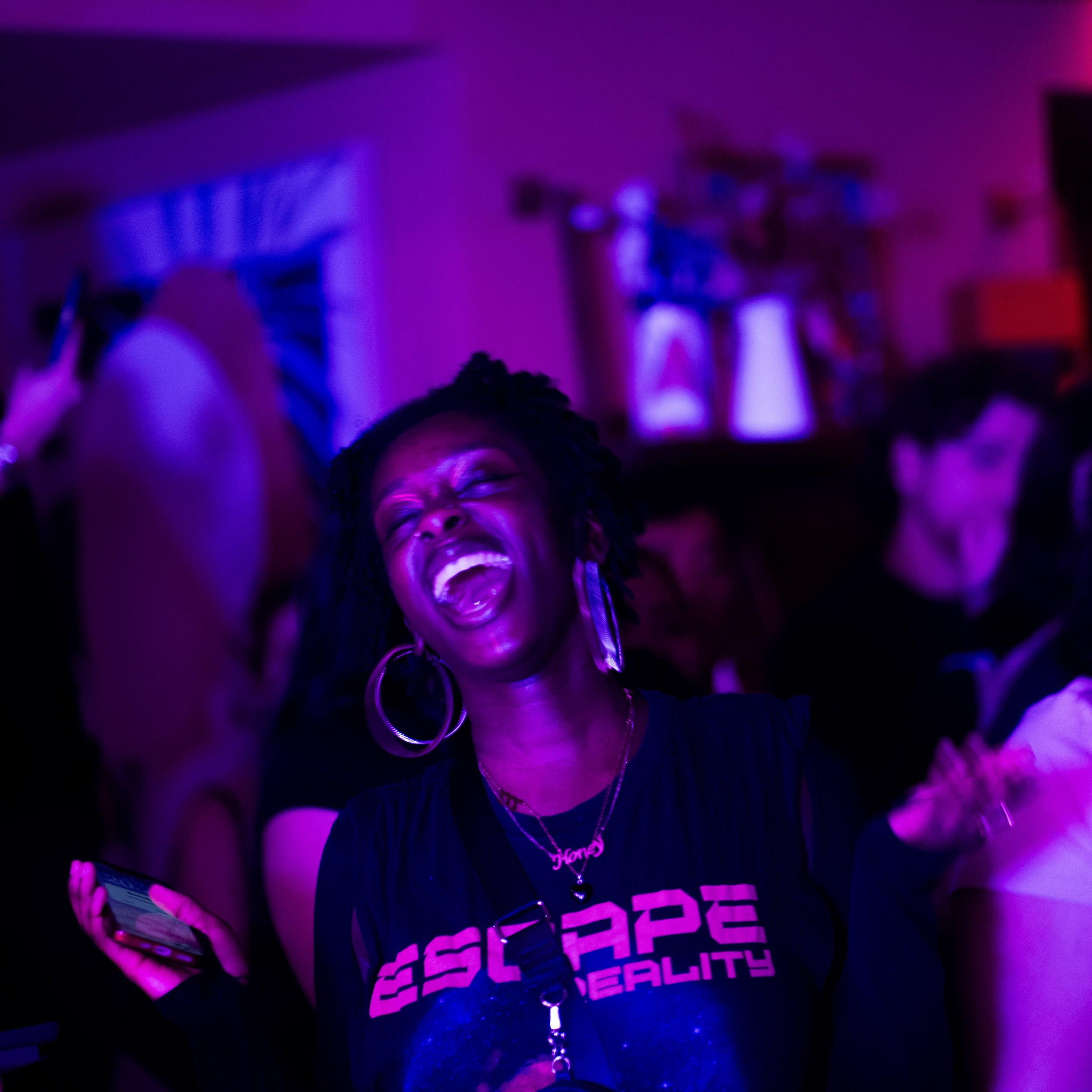 Person at a What Does Not event in London smiling, holding a phone, wearing earrings, a necklace with a heart pendant, and a black T-shirt with pink text. The background is illuminated with pink and purple neon lights.