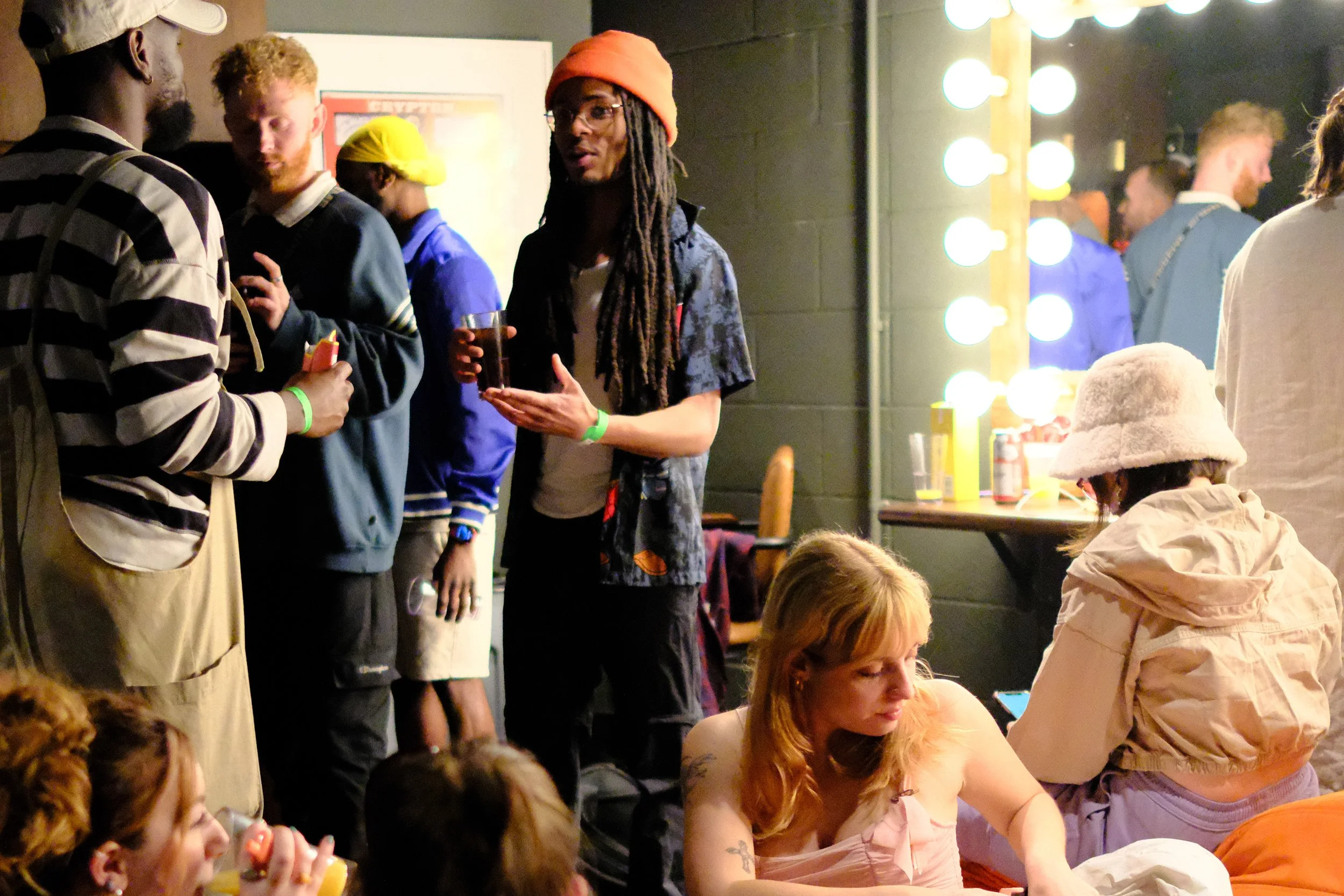 People gathered indoors, some standing and some sitting, with bright vanity lights visible on the right side of the image, in a social or entertainment setting.
