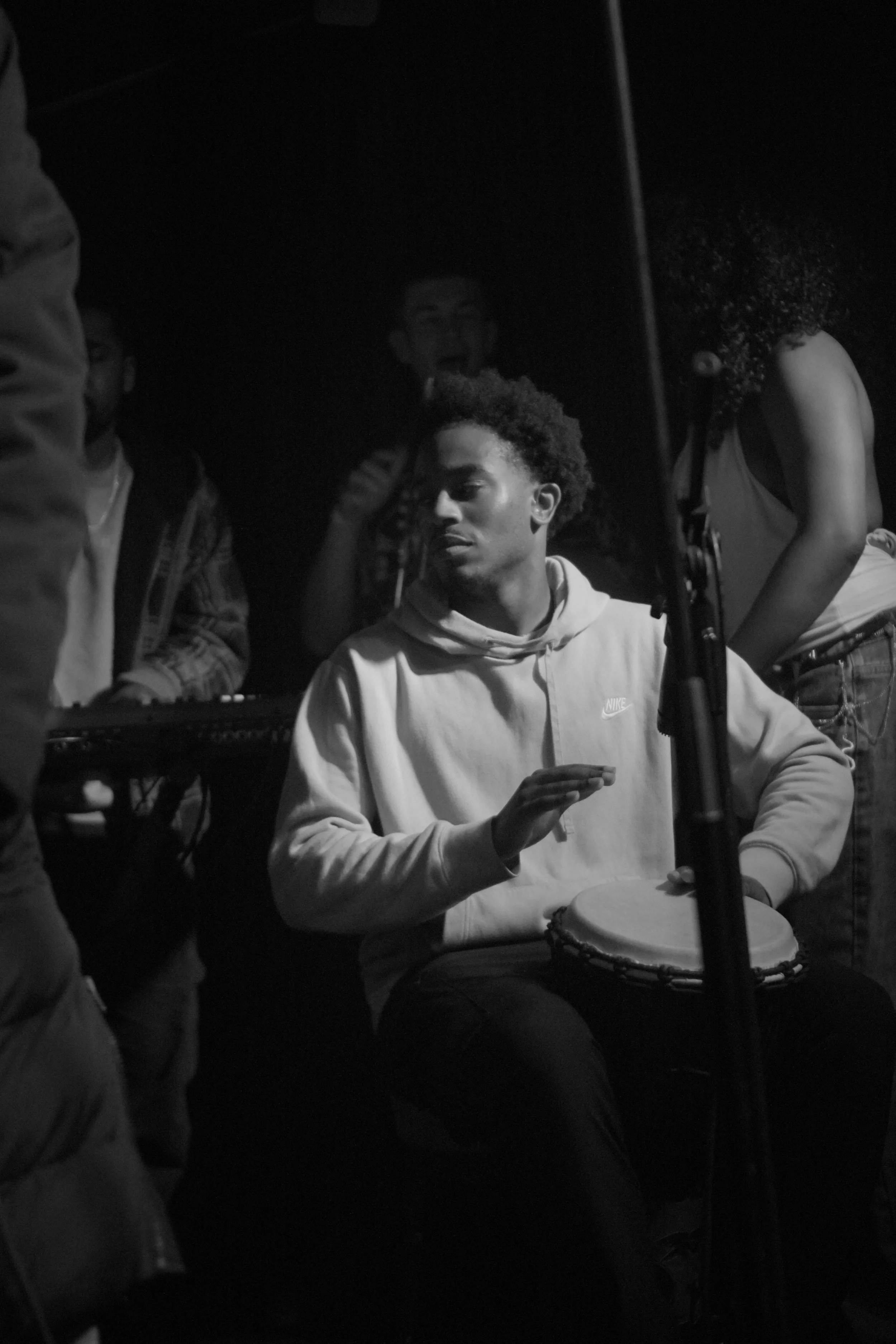 A person with curly hair is playing a drum while others around are engaged, in a dark setting.
