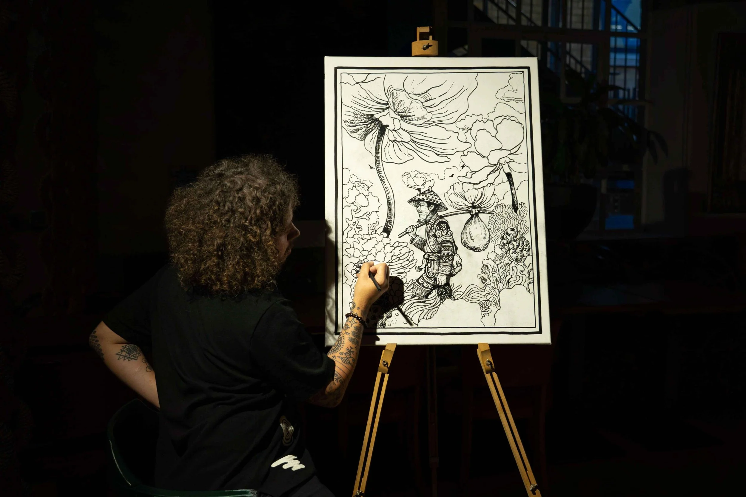 An independent visual artist, a man with tattoos on her left arm and curly hair, dressed in black, is drawing on a black and white artwork featuring a detailed illustration of a fantastical creature, large flowers, clouds & a landscape on an easel.