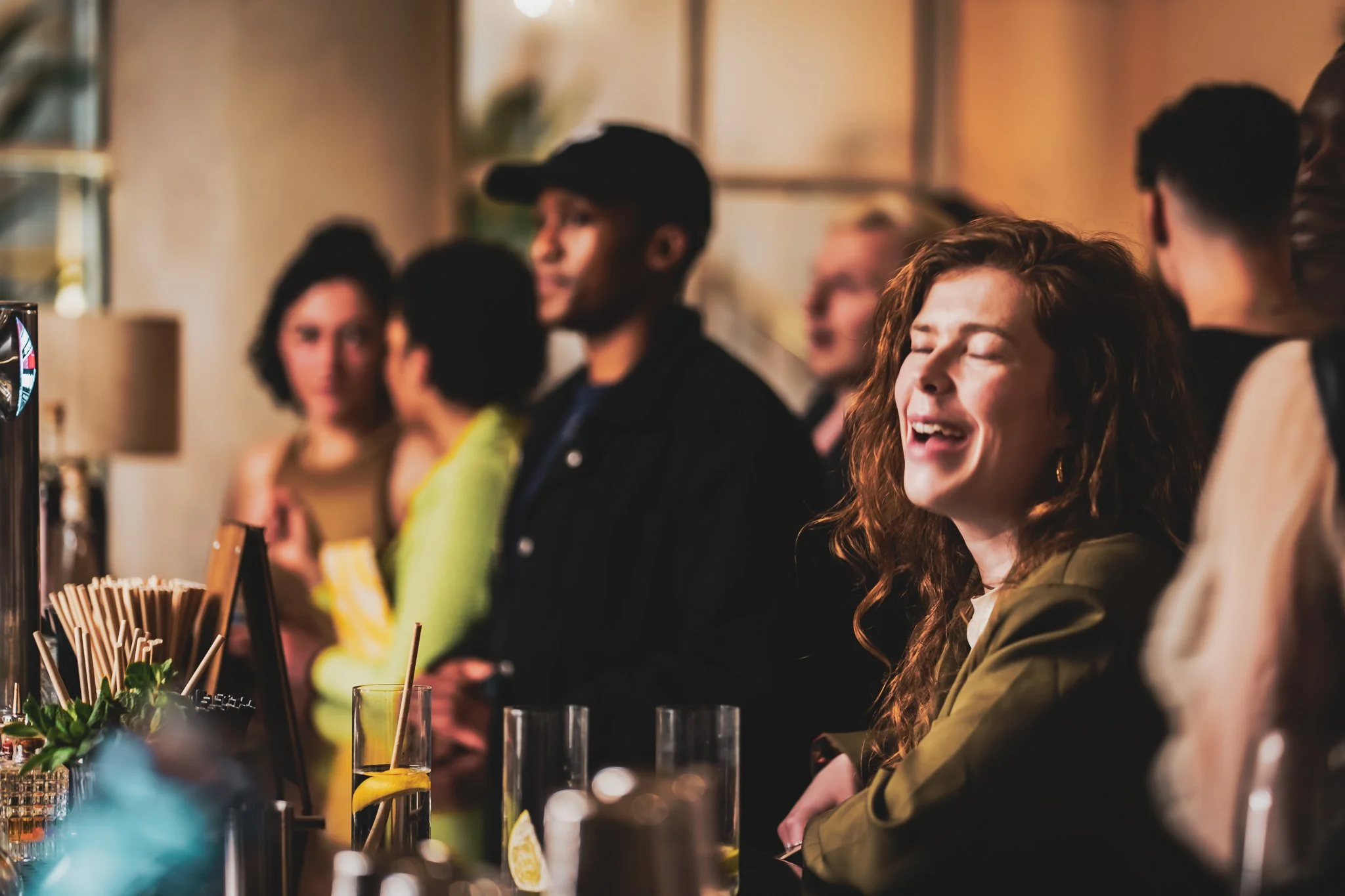 People at a What Does Not creative mixer / social at a bar counter in a dimly lit setting, socializing and enjoying drinks, with a woman in the foreground smiling with eyes closed.