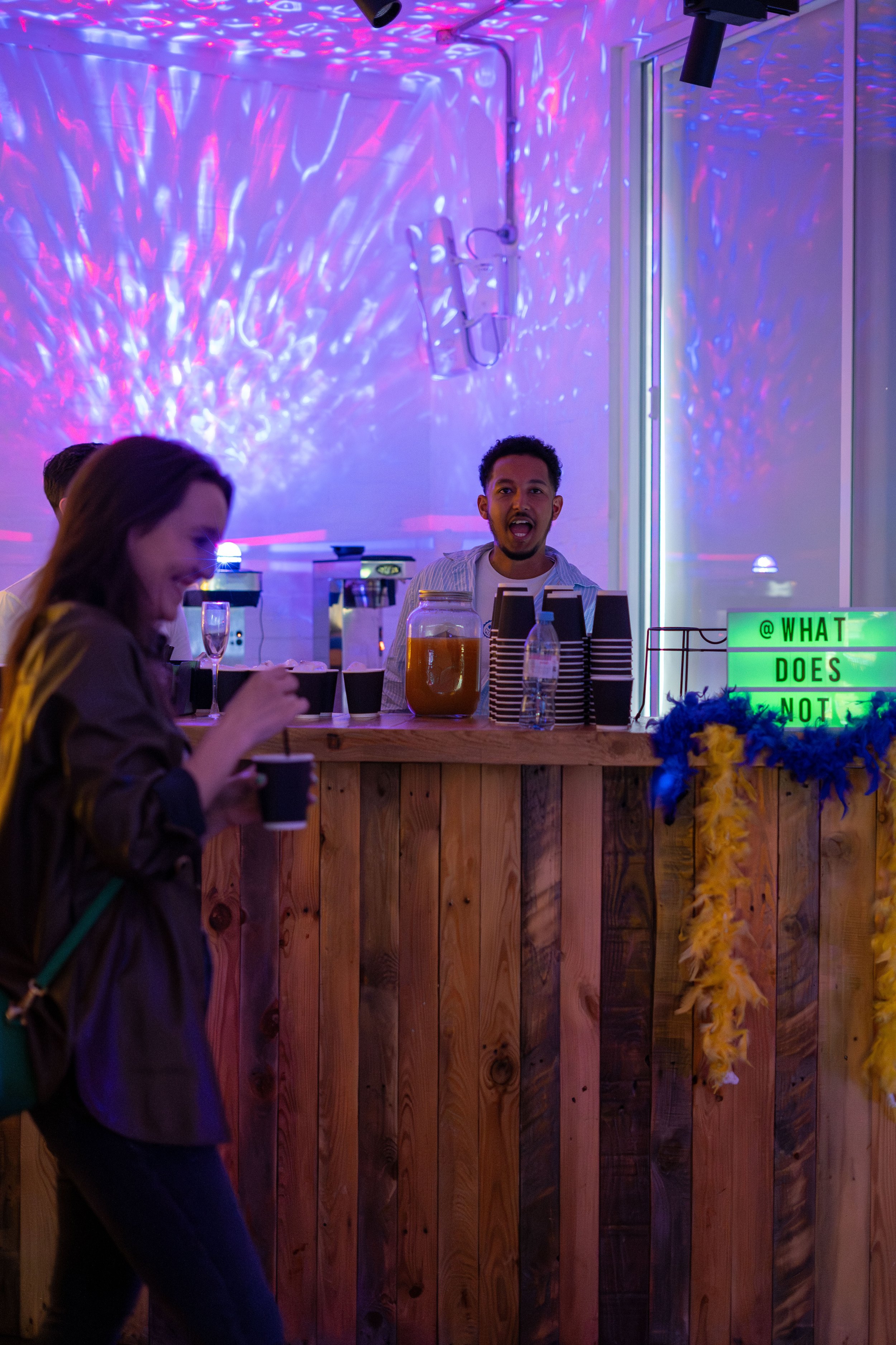 People at a What Does Not house party with purple and pink lighting, standing behind a wooden bar with a sign saying '@WHAT DOES NOT', and a flower arrangement decorated with yellow and blue feathers.