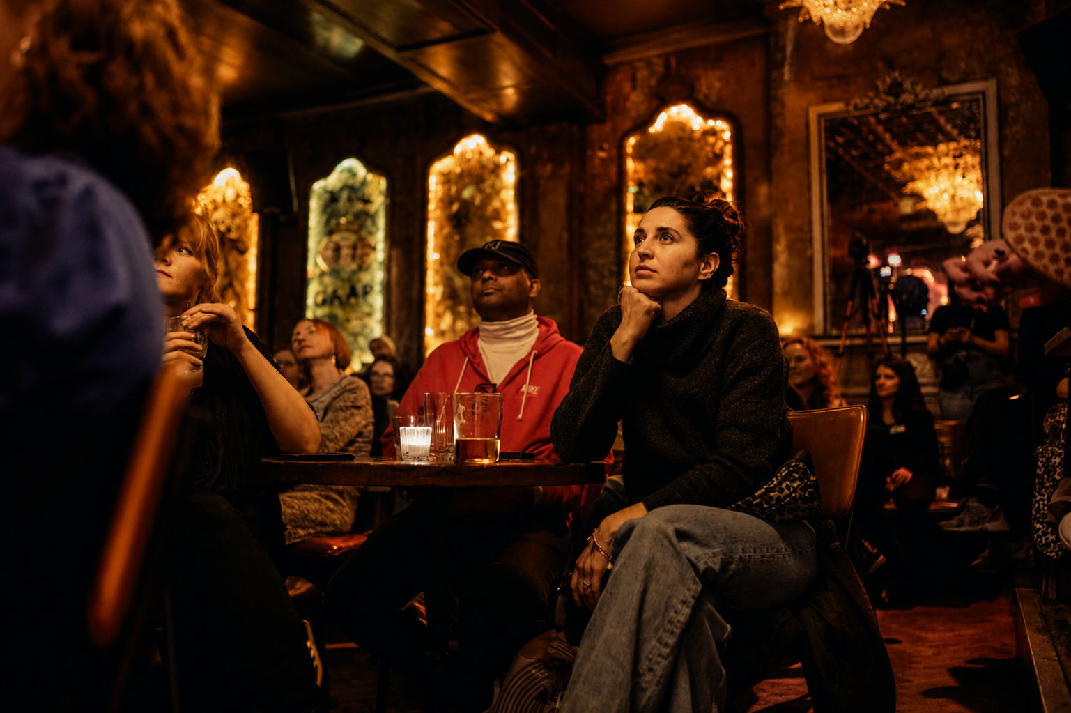 People sitting in a dimly lit lounge, attentively listening to a What Does Not discussion panel. The background features ornate mirrors and warm, golden lighting.