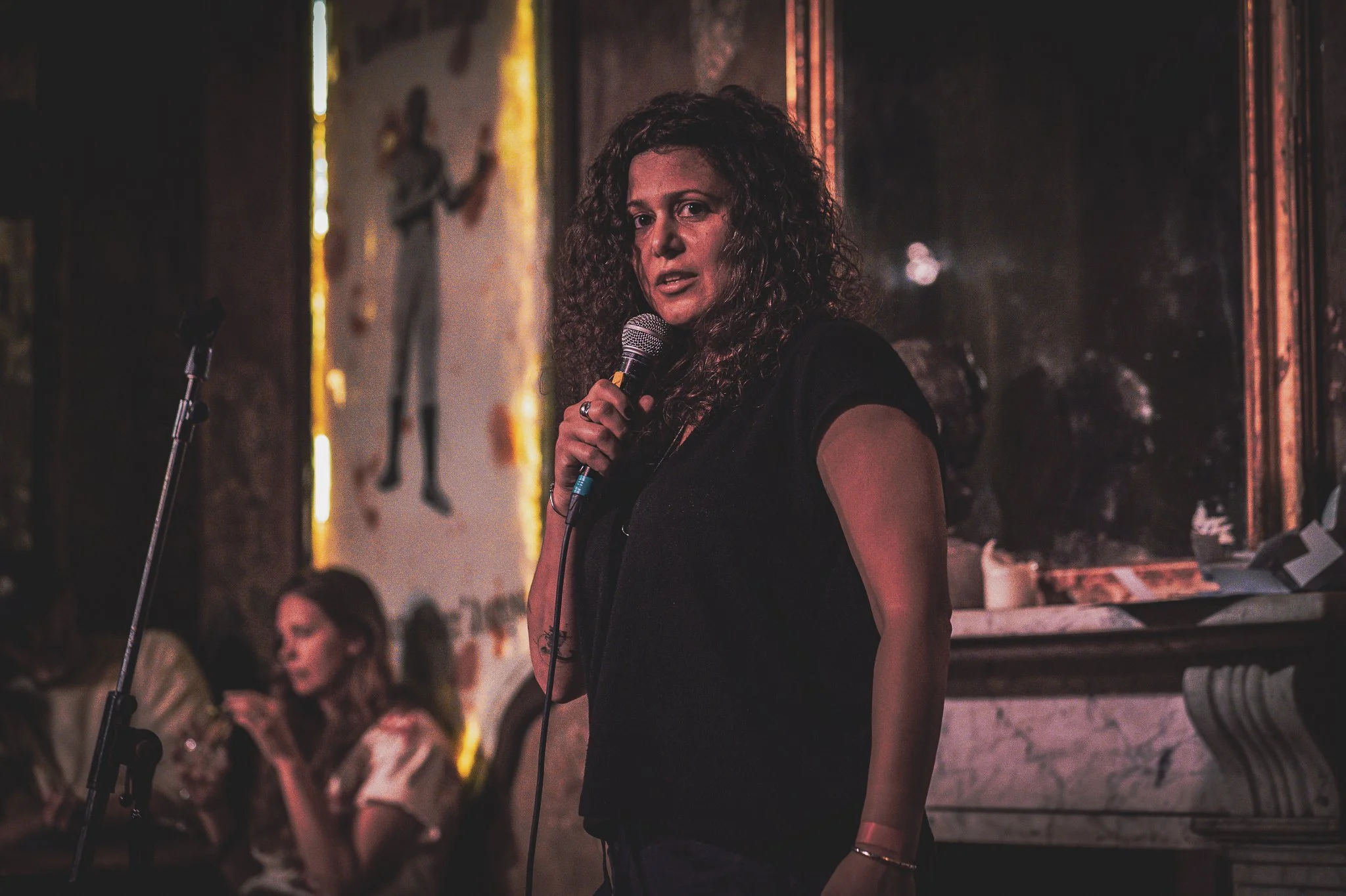 What Does Not founder, Christine Charitonos, a woman with curly hair holding a microphone, performing on stage in front of a seated audience in a dimly lit venue.