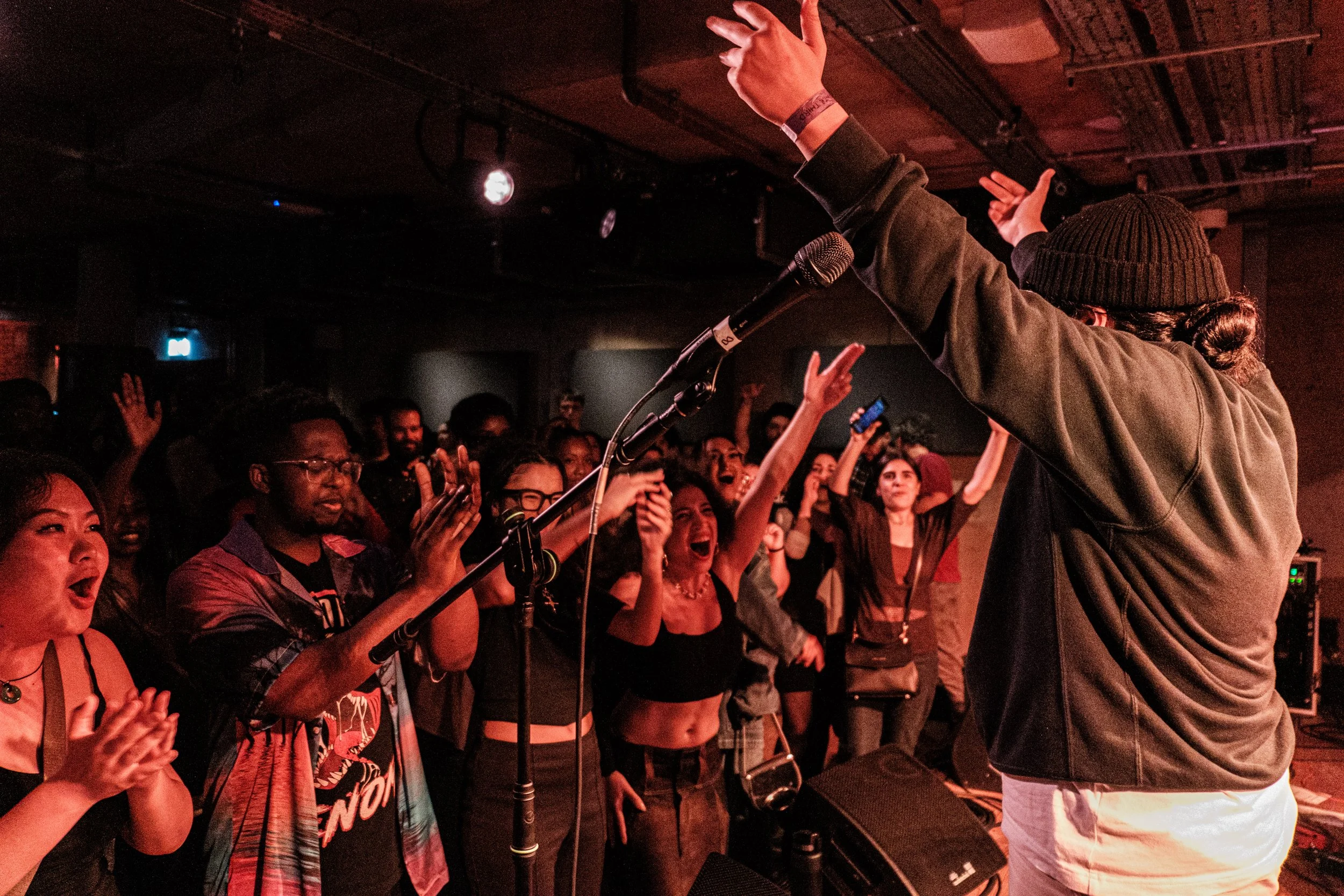 A performer on stage in front of a crowd at a What Does Not event / music showcase for emerging musicians. The person is wearing a beanie and a jacket, gesturing with both hands. The audience is excited, raising their hands and cheering.