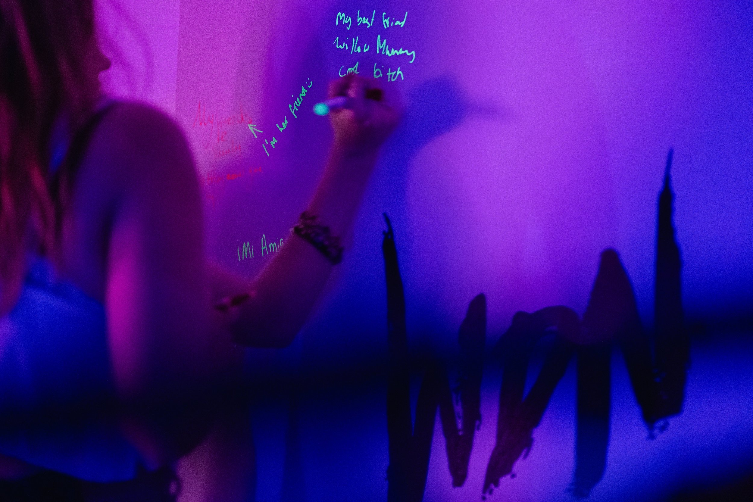 A person writing on an interactive installation at a What Does Not event with neon blue and pink lights. The person's hand is visible, holding a marker.