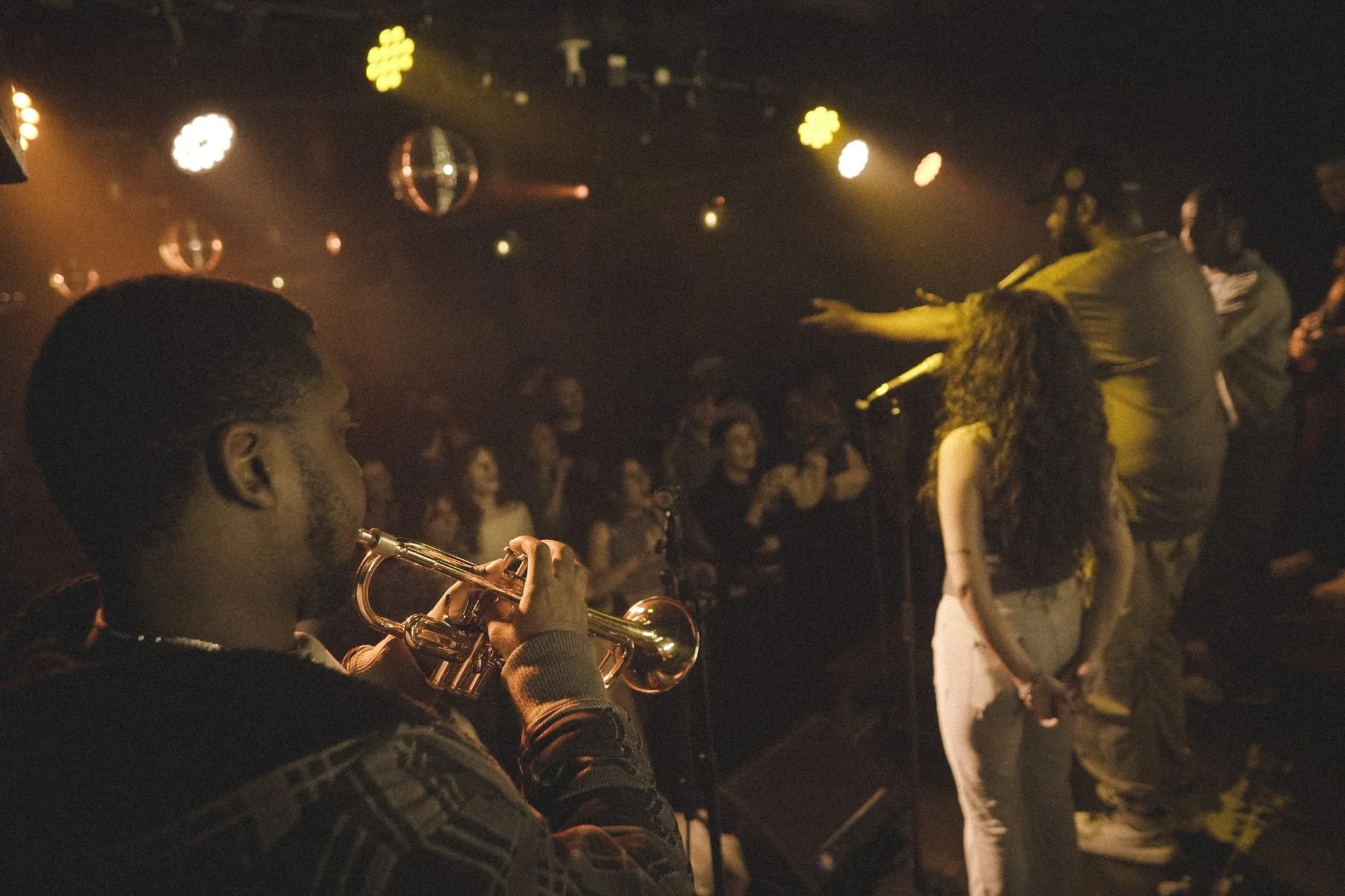 A musician at a What Does Not Afternoise open jam, music improv night, playing trumpet in a dark, crowded music venue with disco balls and stage lights, and an audience in the background.