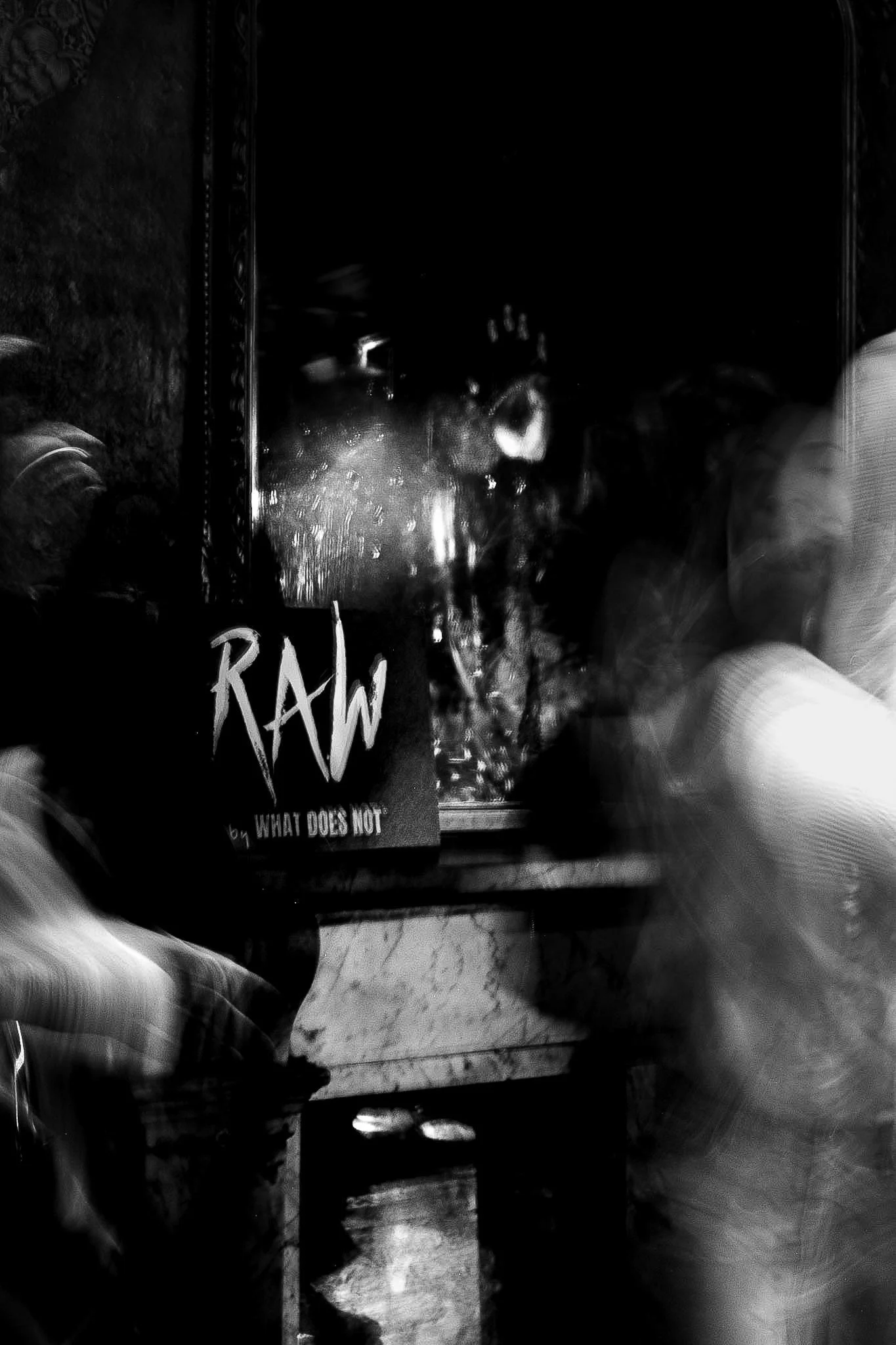 An artistic shot from a RAW uncensored storytelling night featuring a dark, monochrome photo of a mirror reflecting a person with long hair, a hand raised near the face, and a makeup brush, with a partially visible book titled 'RAW' on the table.