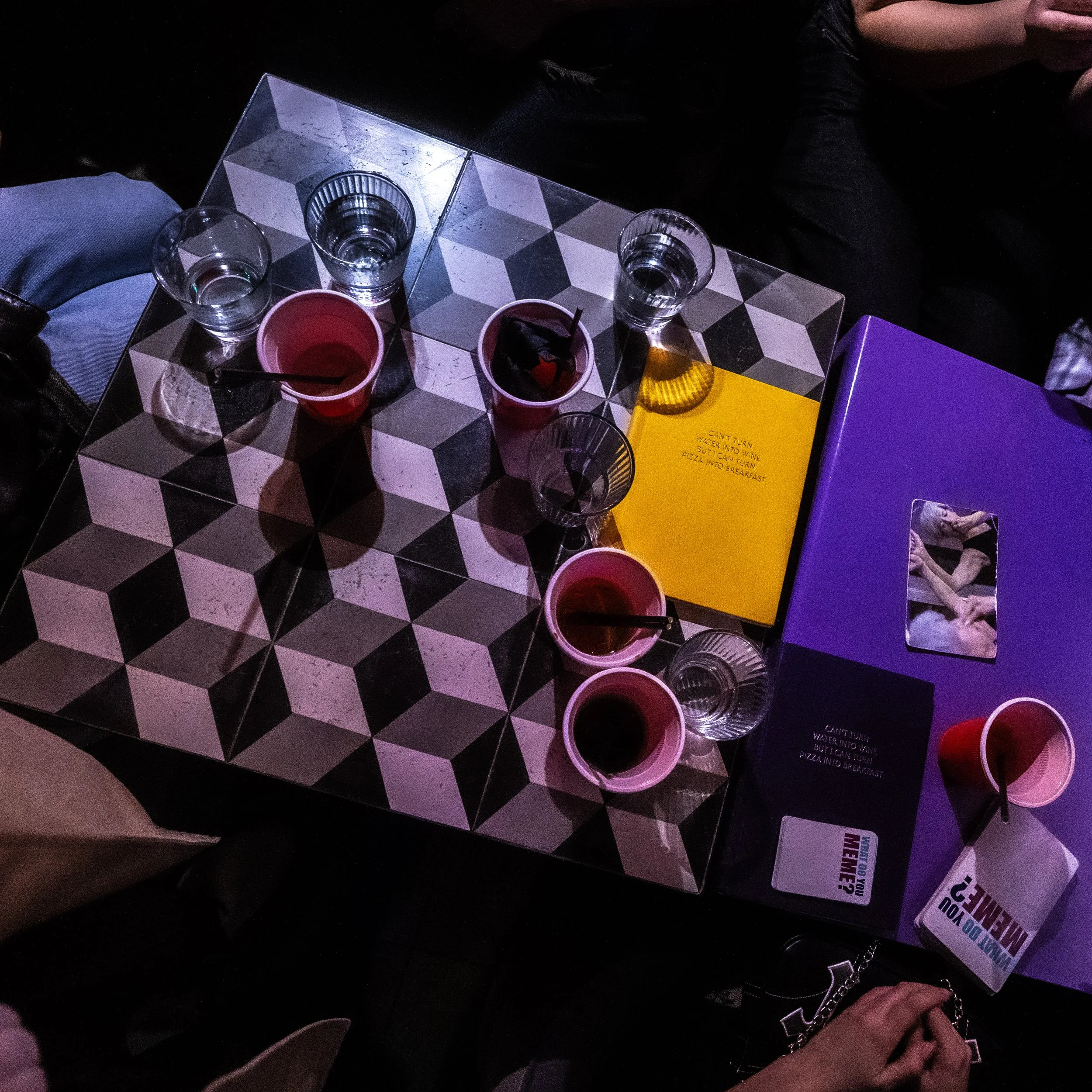 A table with seven glasses of water, two red cups, a yellow menu, and a magazine with a photo of a woman and a dog, in a dimly lit setting.