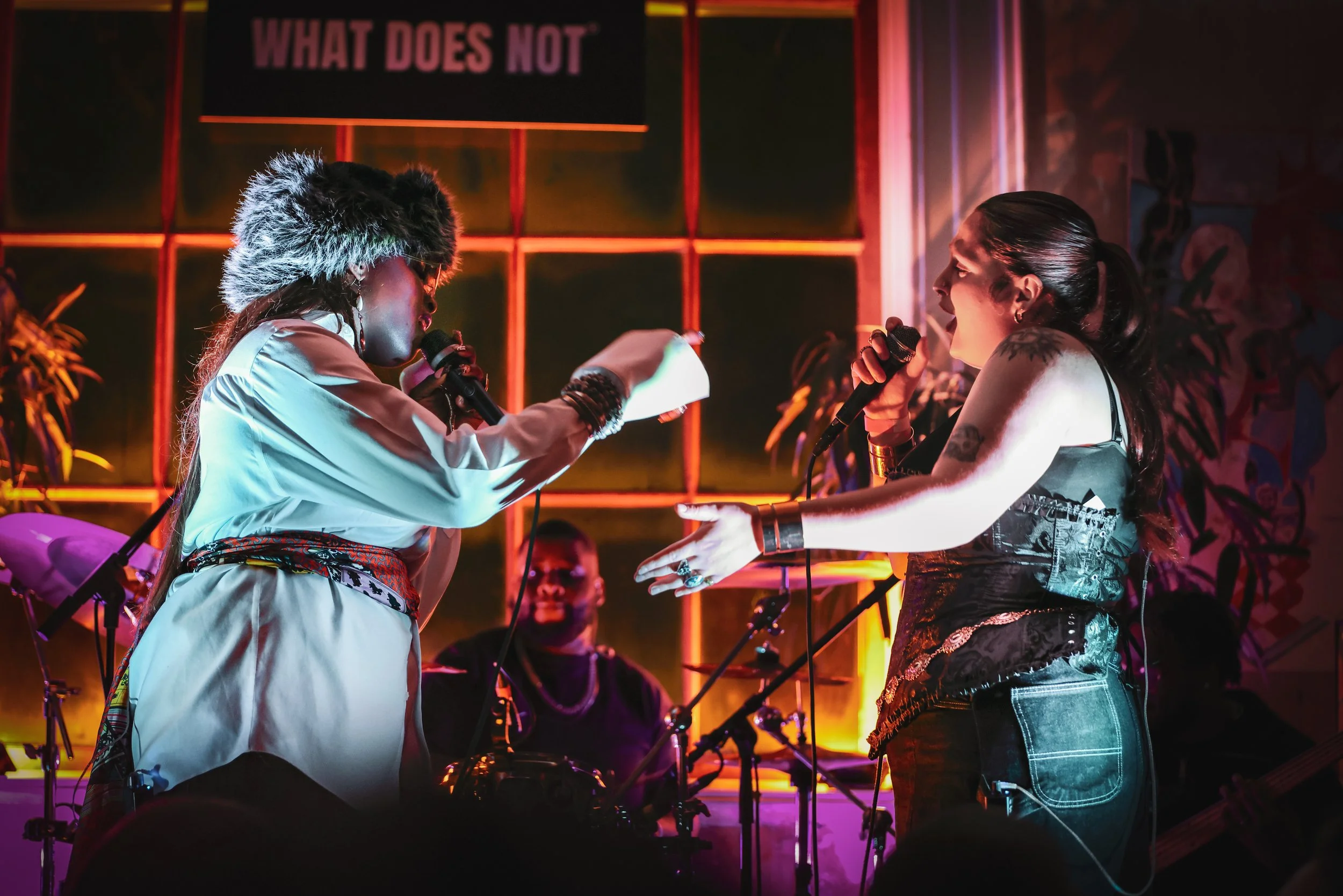 Two emerging musicians sing on stage as part of a headline show produced by What Does Not at a live music night in London, singing into microphones, with a sign behind them that reads "What does not" and a band playing in the background.