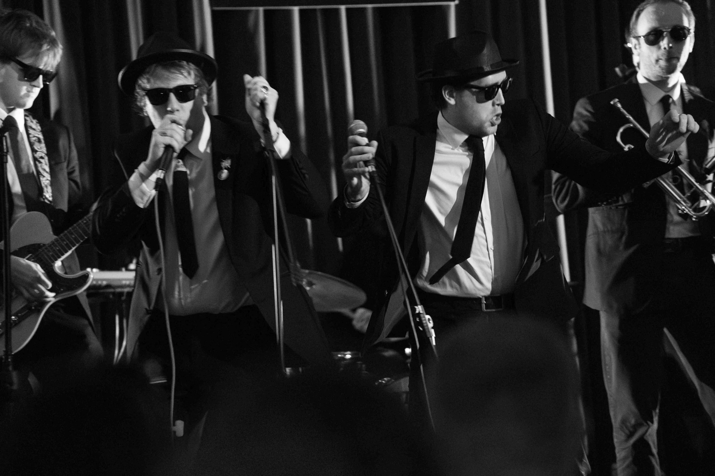 A black-and-white photo of a musical performance featuring five band members dressed in suits, wearing sunglasses, and playing instruments including drums, guitar, microphone, and trumpet.