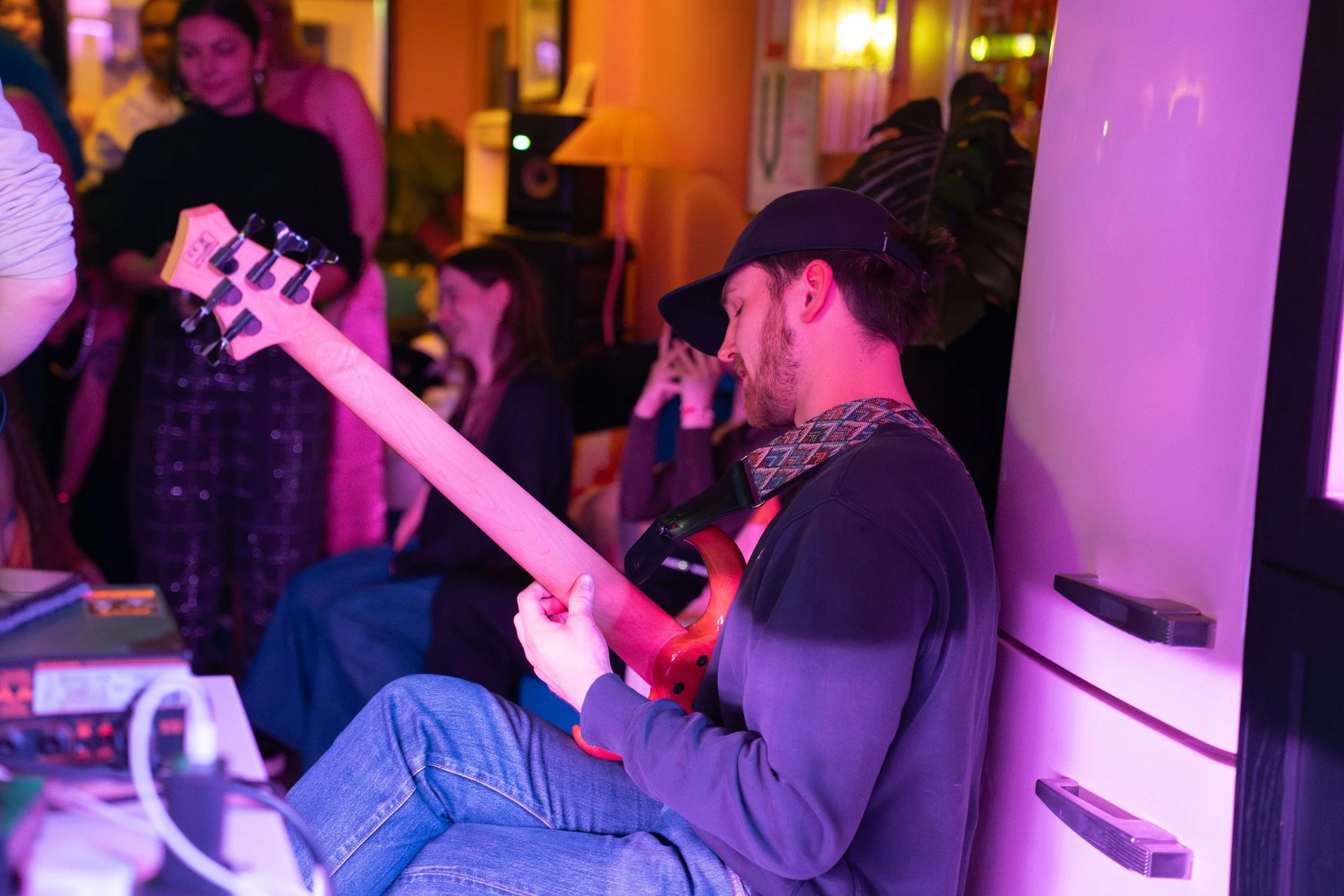 An emerging musician in London seated, wearing a black baseball cap playing a bass at a What Does Not open jam, with two women in the background in a room with colorful lighting.