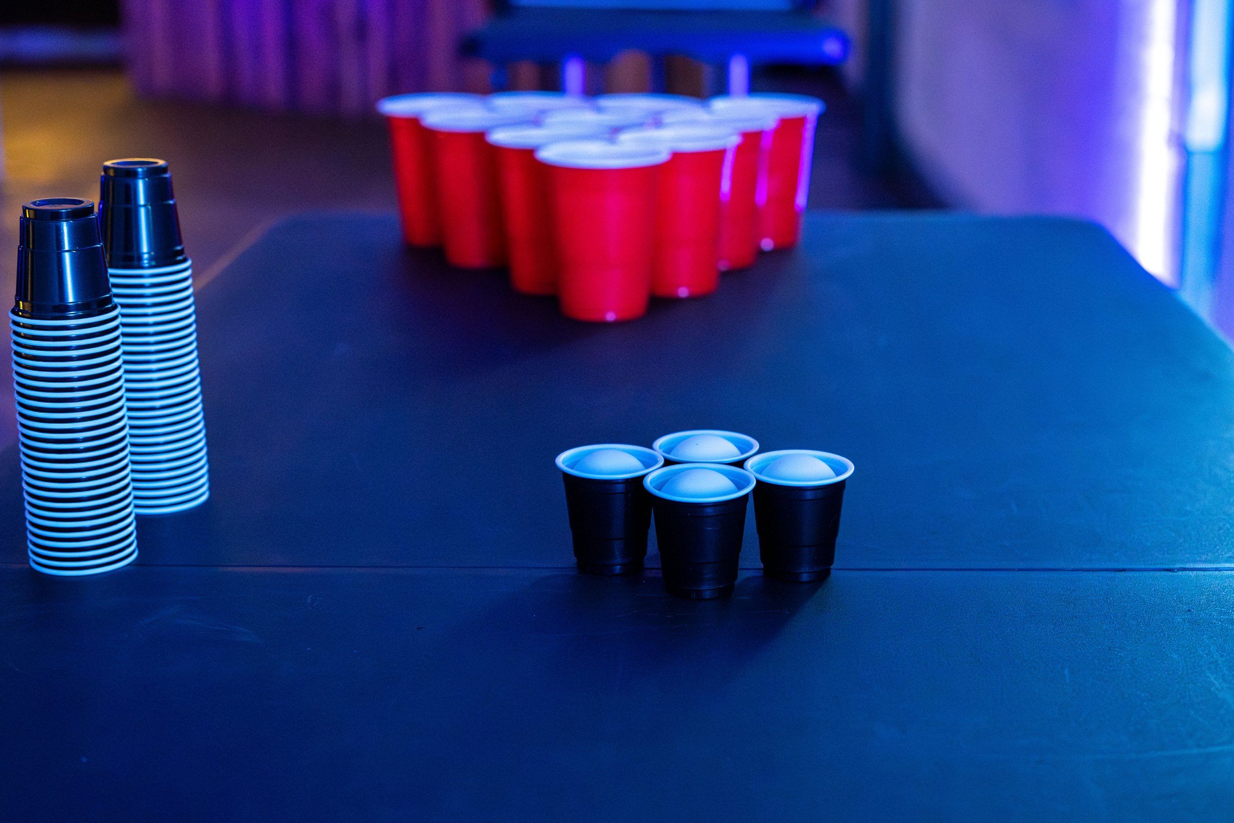 Red plastic cups arranged in a triangle, black plastic cups arranged in a triangle to play beer pong at a What Does Not house party, and stacks of white disposable cups on a black table, with a dark background illuminated by blue and purple lighting