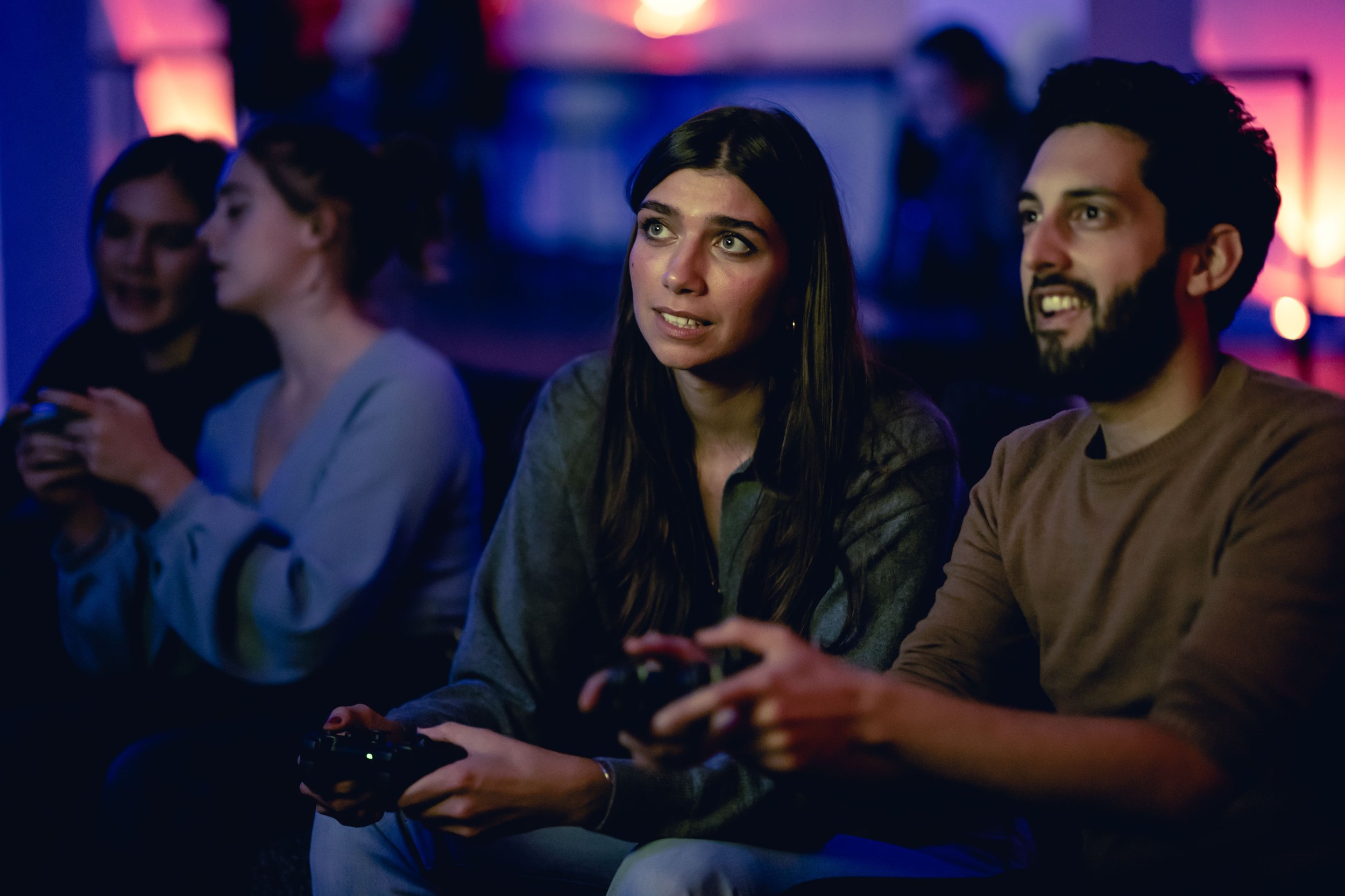 Four people sitting in a dark room with colorful lights, one woman with long dark hair holding a game controller, looking engaged, others looking in different directions, some smiling or talking as they play games at a What Does Not house party