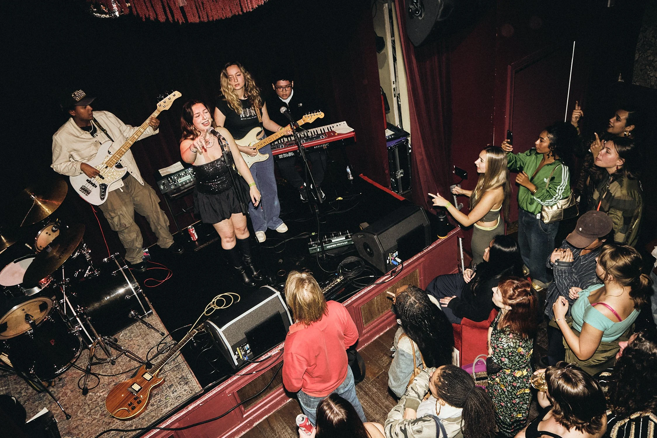 A live band performs on stage at a small venue with an audience standing close at a live music night in London by What Does Not. The band includes a female vocalist, a guitarist, a bassist, and a keyboard player.