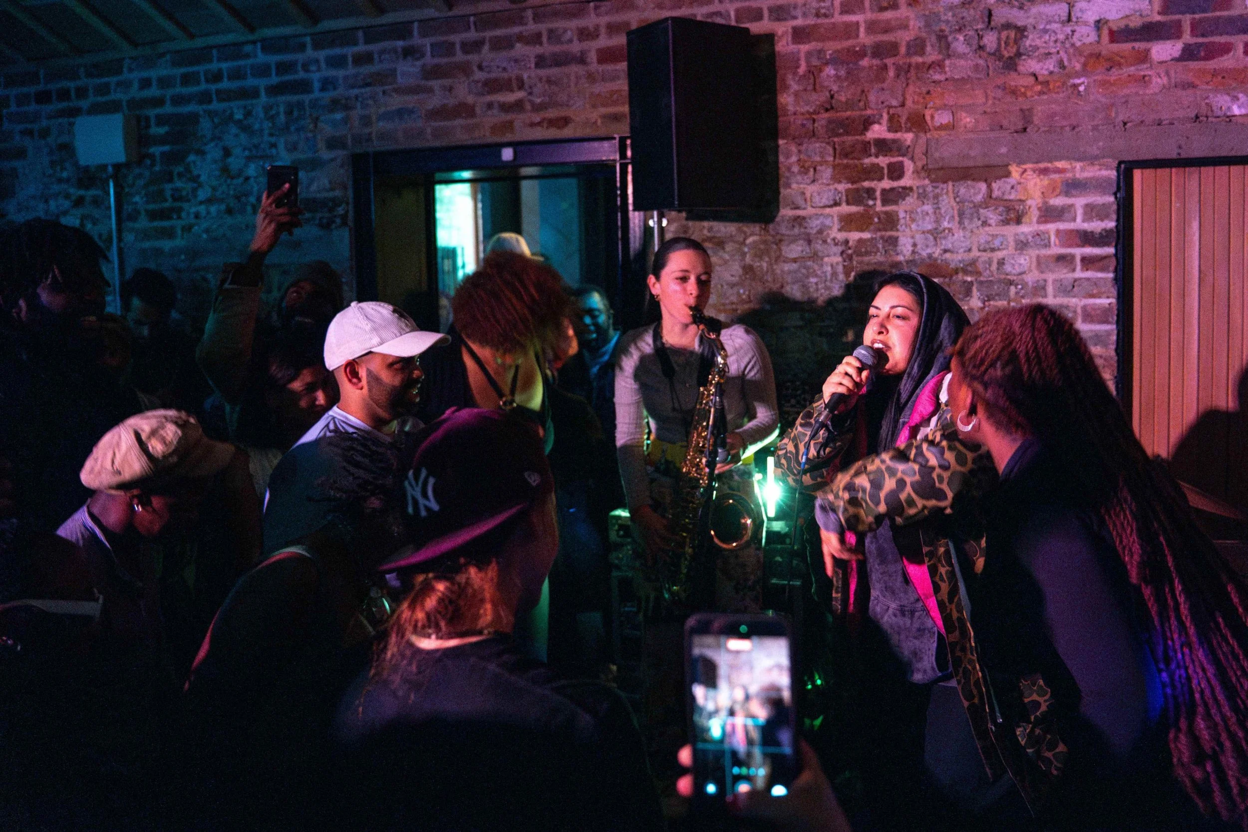 A group of people at a bar, with some performing music, including a woman singing into a microphone and a woman playing a saxophone, with dim lighting and a brick wall background, at a What Does Not open jam session in London