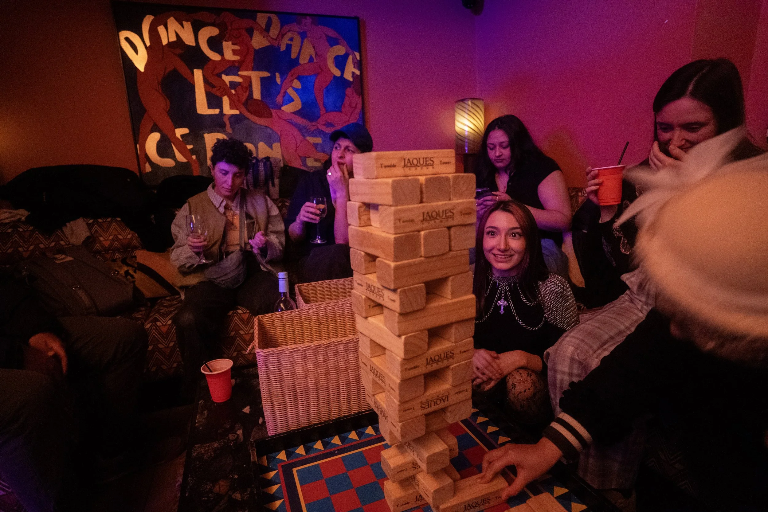 A group of people playing a large outdoor Jenga game at a What Does Not house party in London. The Jenga tower is tall with wooden blocks, and the people look engaged and enjoying themselves