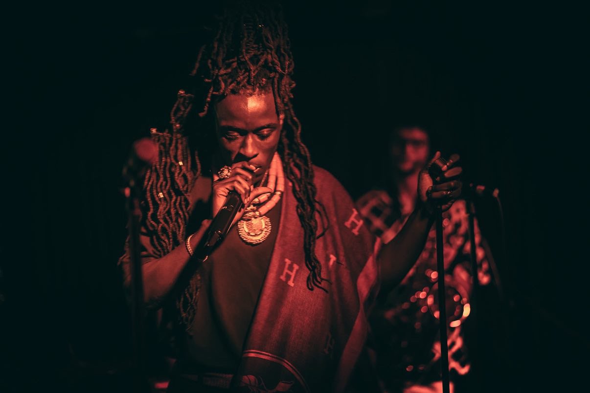 An independent artist and musician in London with long dreadlocks sings into a microphone on a dark stage, wearing a red patterned garment and jewelry, as part of a What Does Not headline live music show in London