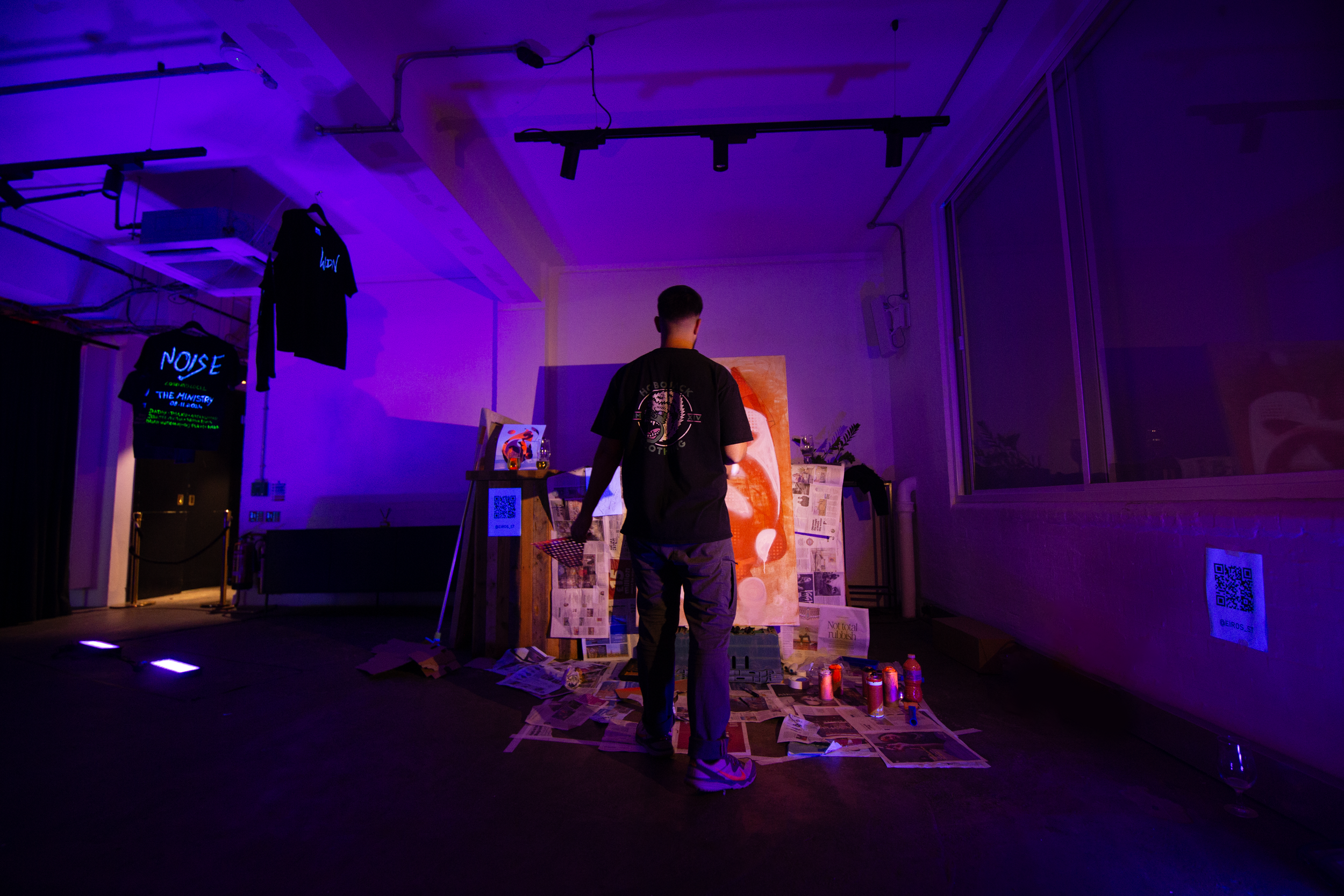 Person standing in a studio like setting at a RAW uncensored storytelling in London with purple lighting, facing a large canvas on an easel surrounded by newspapers and spray paint cans.