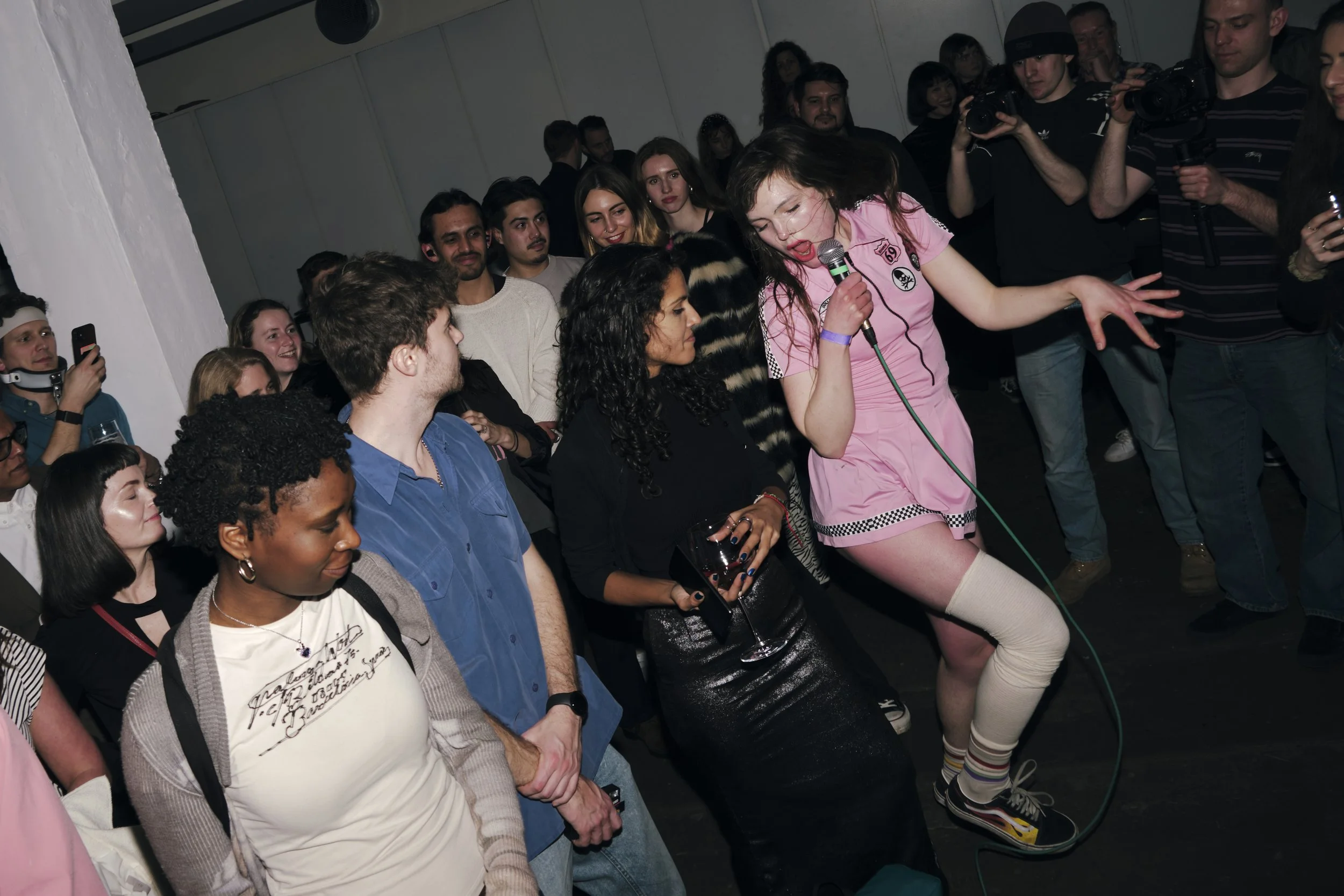 A woman wearing a pink dress sings into a microphone at a crowded What Does Not event in London with an audience watching and taking photos.