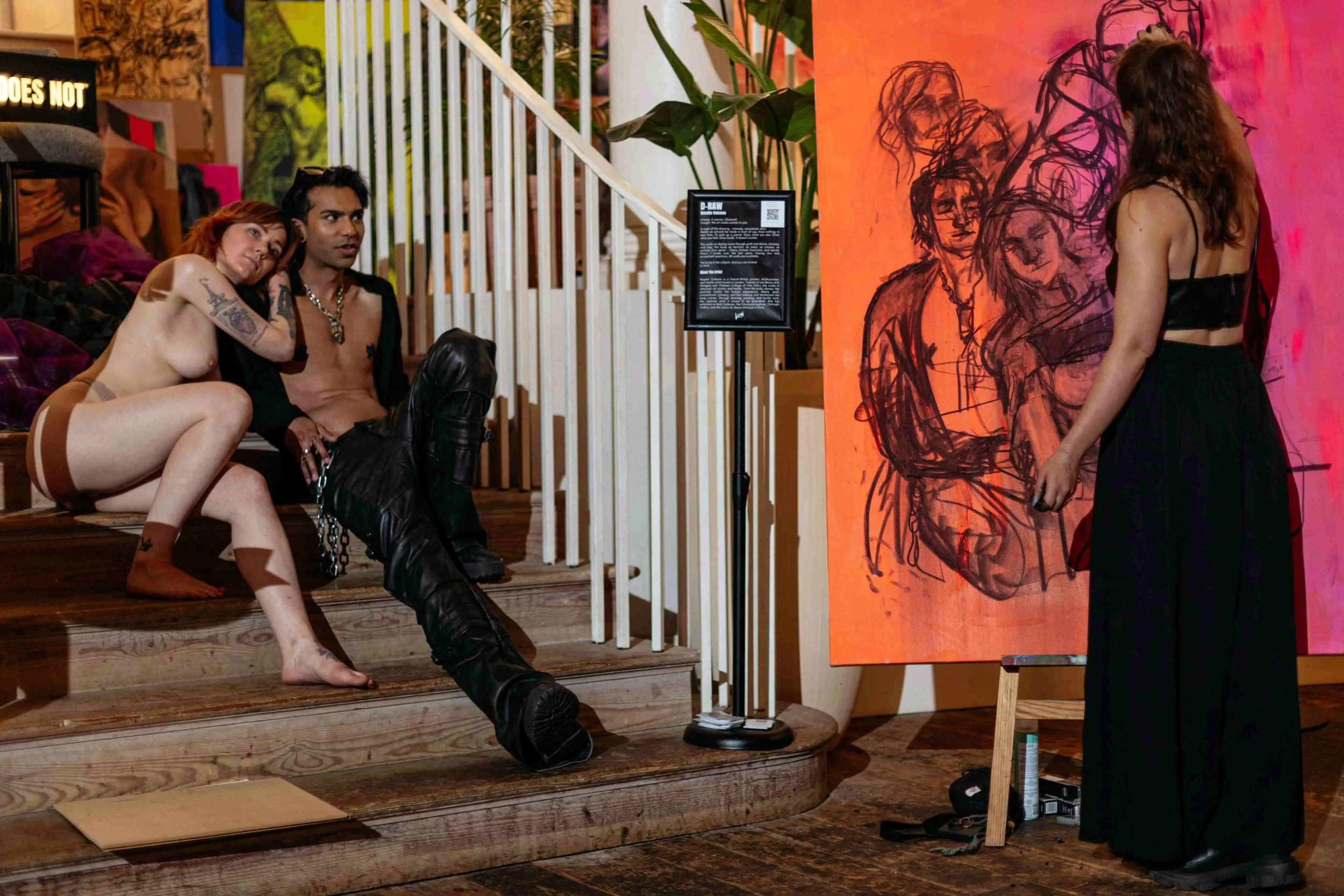 Two people sitting on wooden stairs near an art exhibition, with a woman in black dress standing near a painting of sketched faces on a red-orange background.