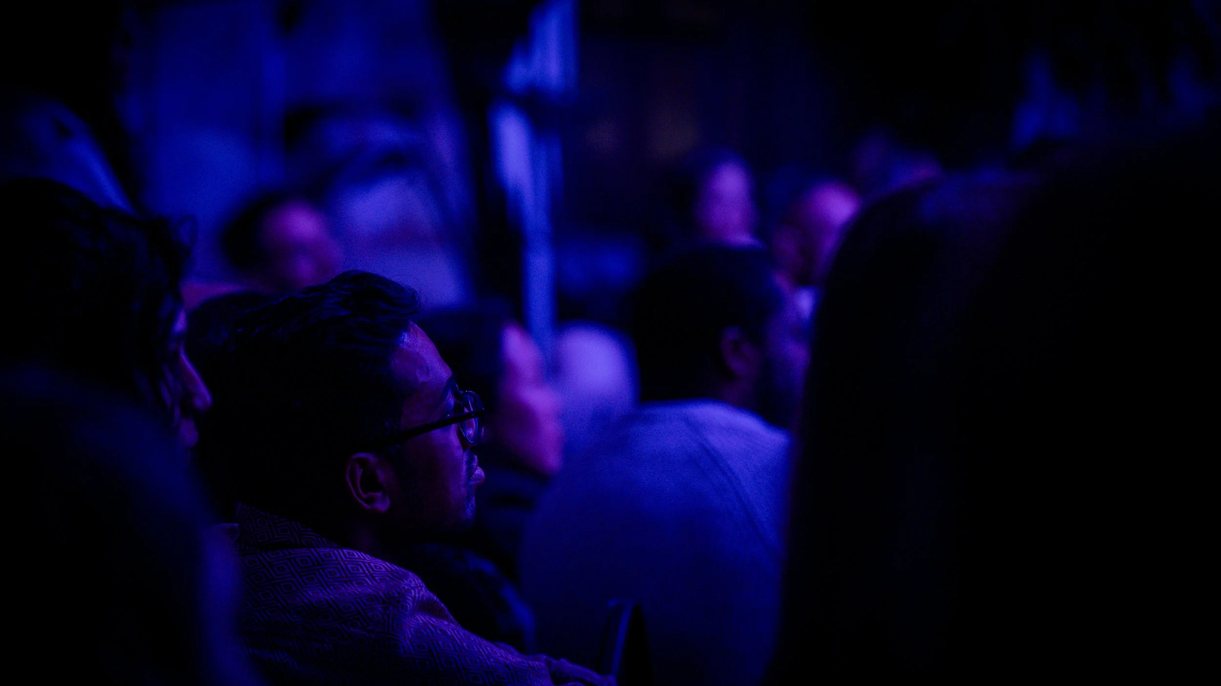 People sitting in an audience under purple lighting, watching the storytellers at RAW share their stories.