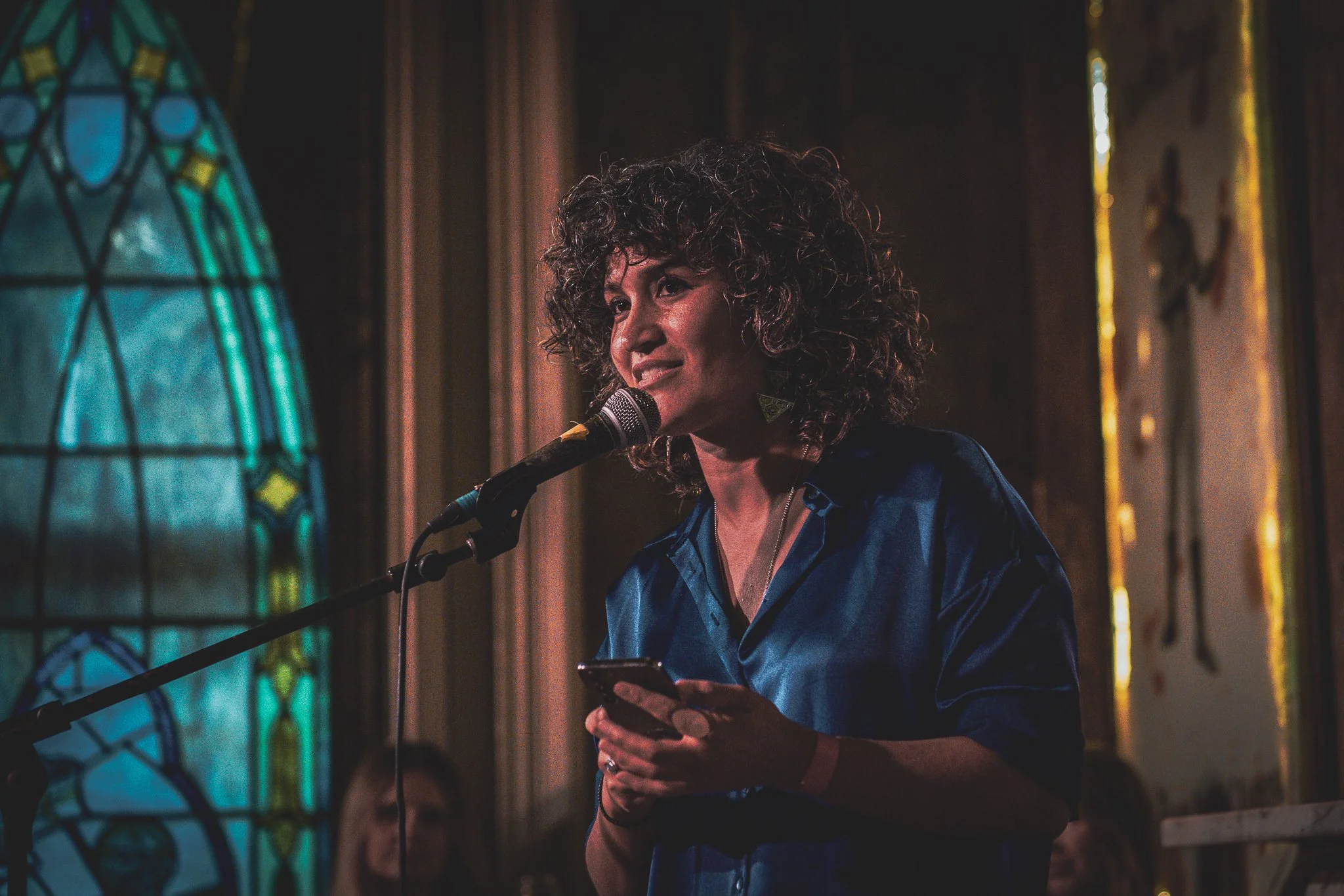 What Does Not head of partnerships and content, Mary Valiakas, a woman with curly hair holding a smartphone and speaking into a microphone at a RAW uncensored storytelling night.
