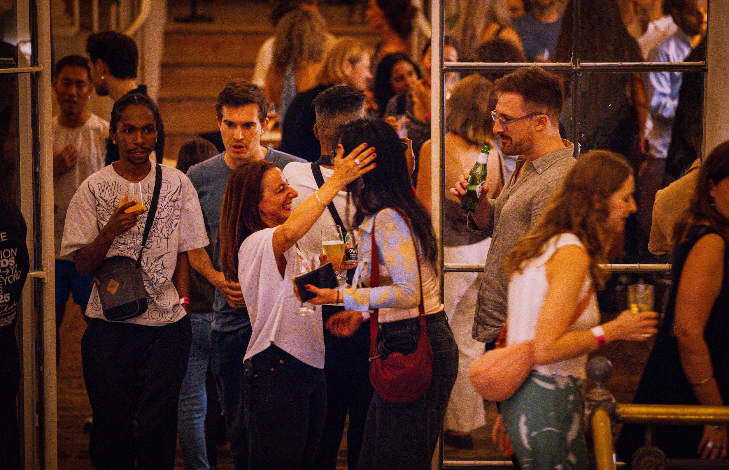 A group of diverse people at a What Does Not social / creative mixer, standing near a glass door with some holding drinks, engaging in conversation.