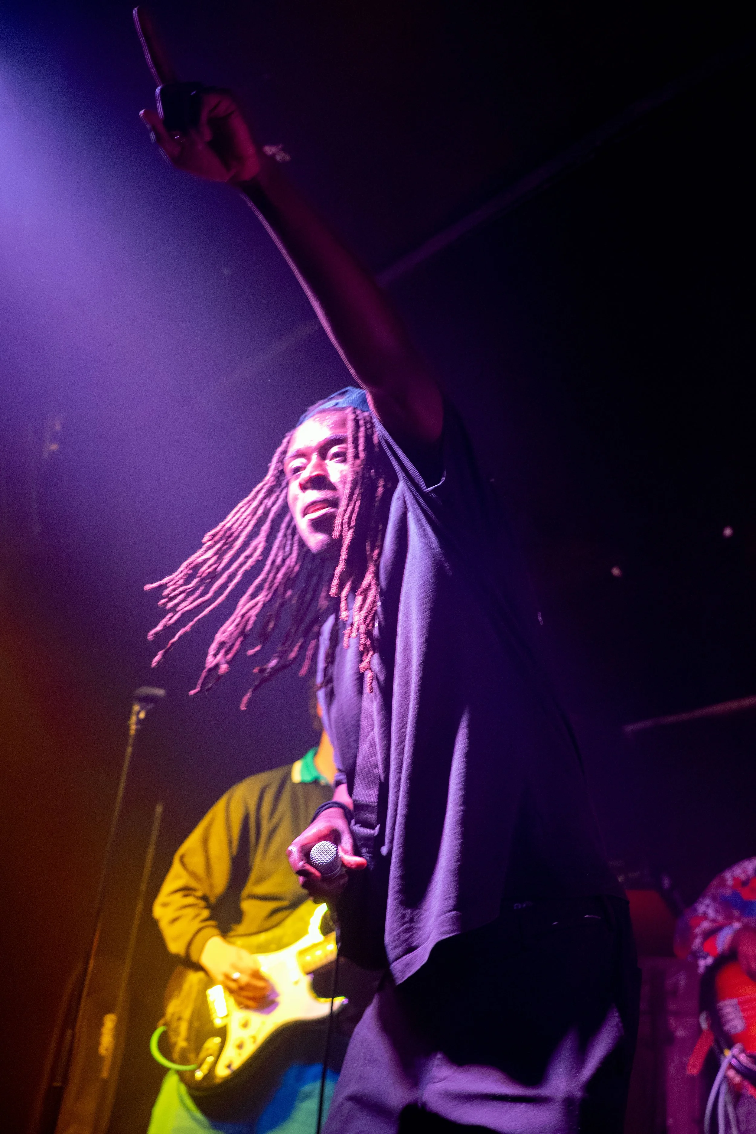 A musician with long dreadlocks holding a microphone on stage with colorful lighting, a guitarist in a yellow jacket playing guitar in the background, and another person partially visible on the right.