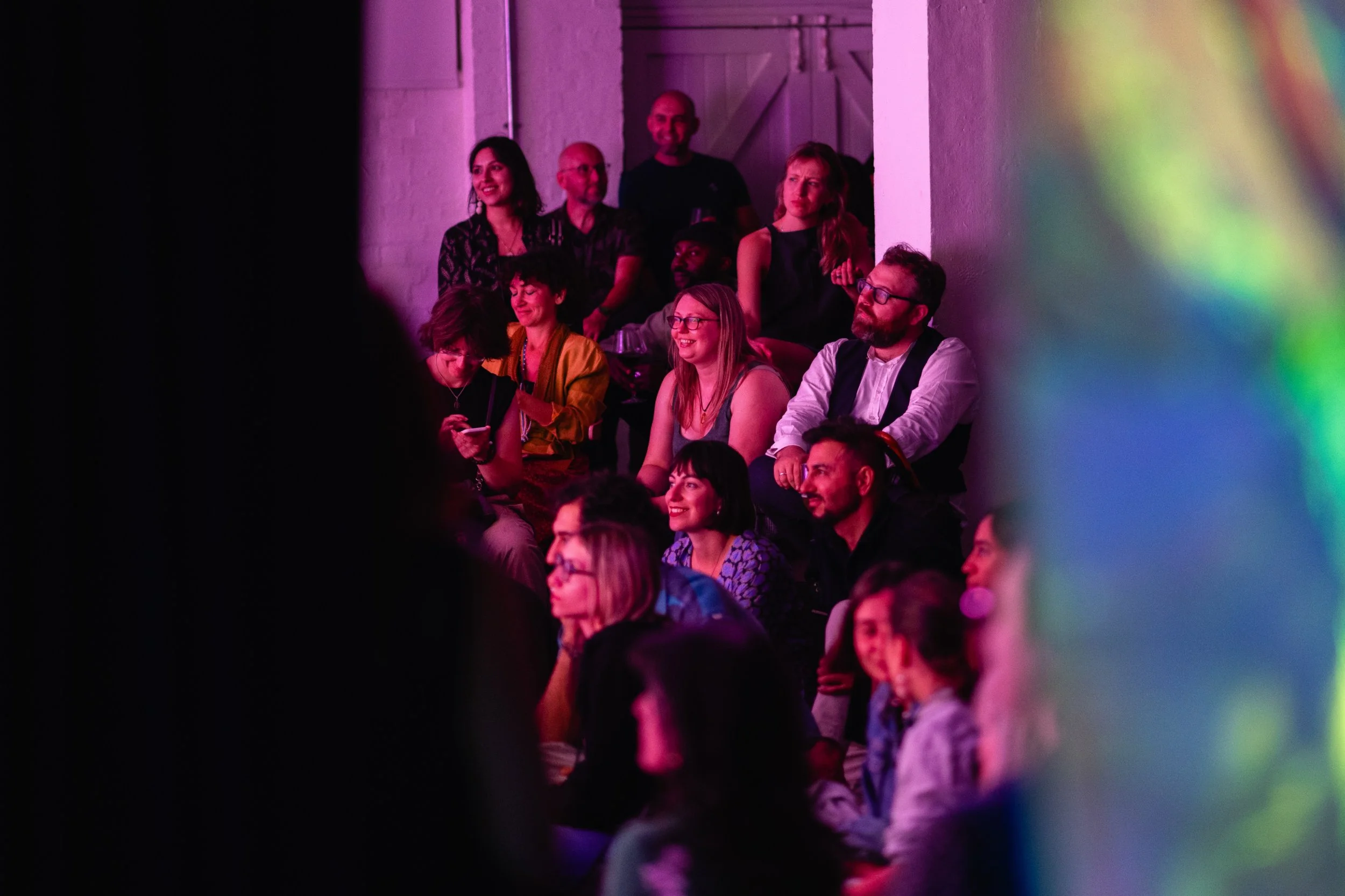 Group of people standing and sitting in a dimly lit room at a RAW uncensored storytelling night, some smiling and others looking serious, with colorful art in the foreground
