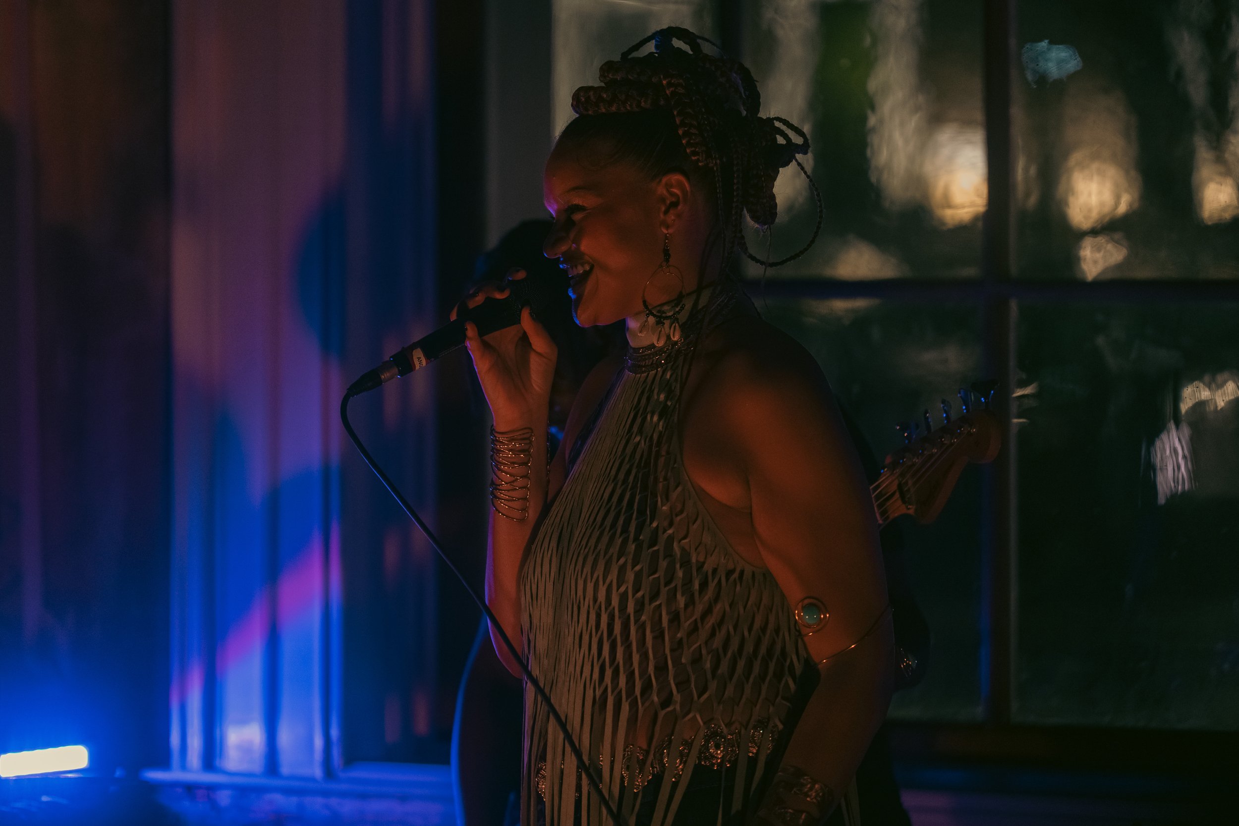 An emerging musician and independent artist, a woman, sings into a microphone in a dimly lit room with vibrant blue, purple, and yellow lighting.