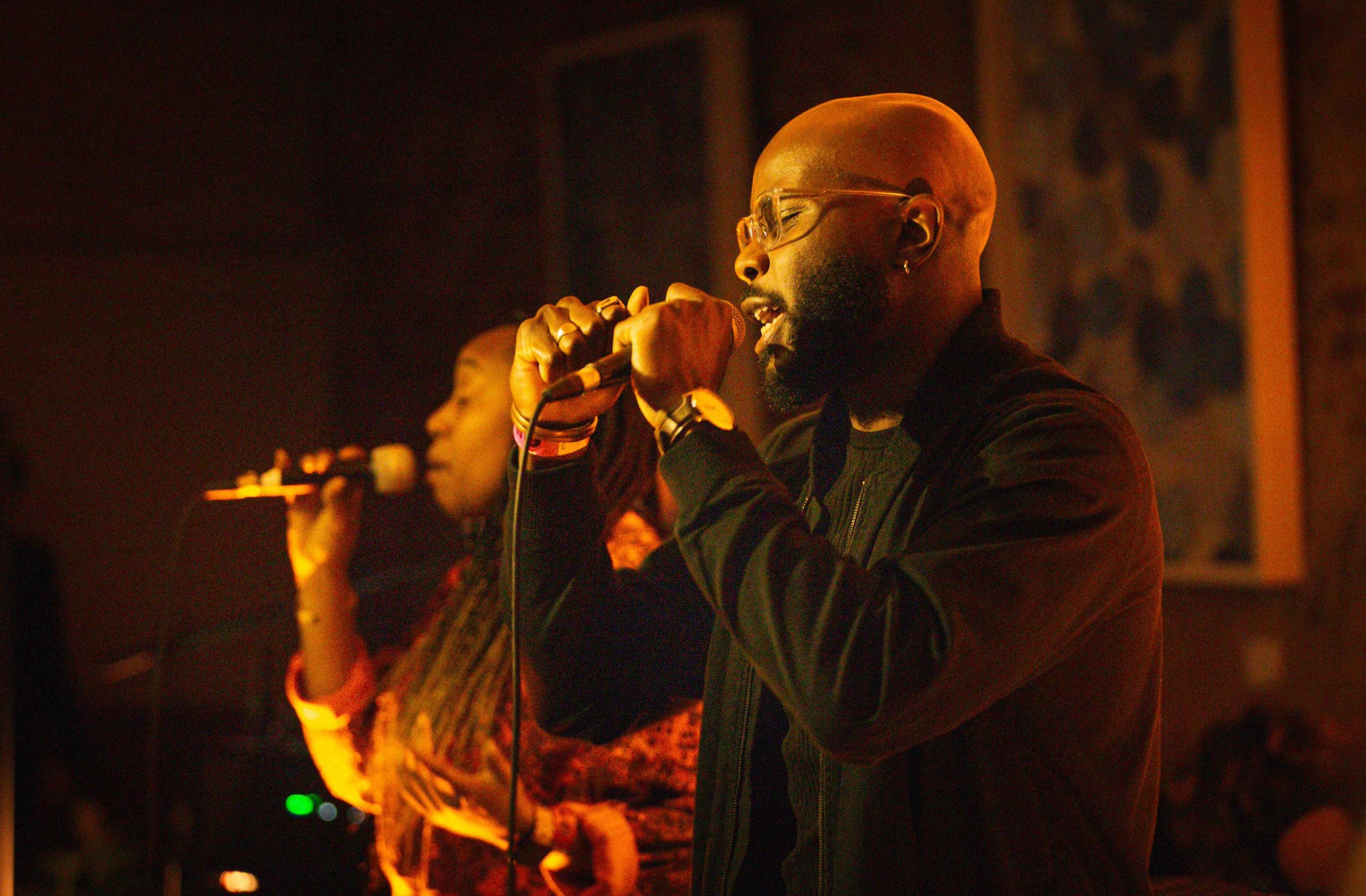 A man with glasses and a beard singing into a microphone in a dimly lit setting.