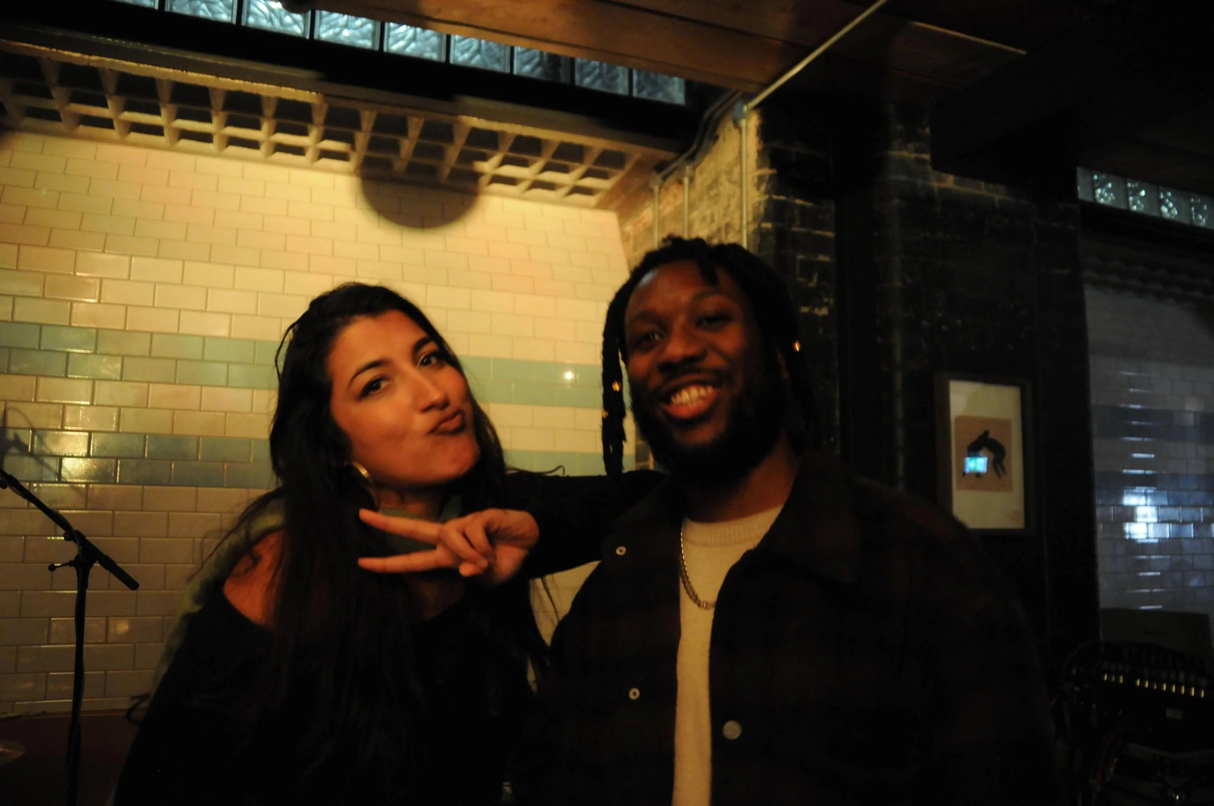 Two people posing for a photo indoors against a brick wall. The woman on the left is making a peace sign with her hand, has long dark hair, and is smiling. The man on the right has long dreadlocks, is smiling widely, and is wearing a dark jacket over
