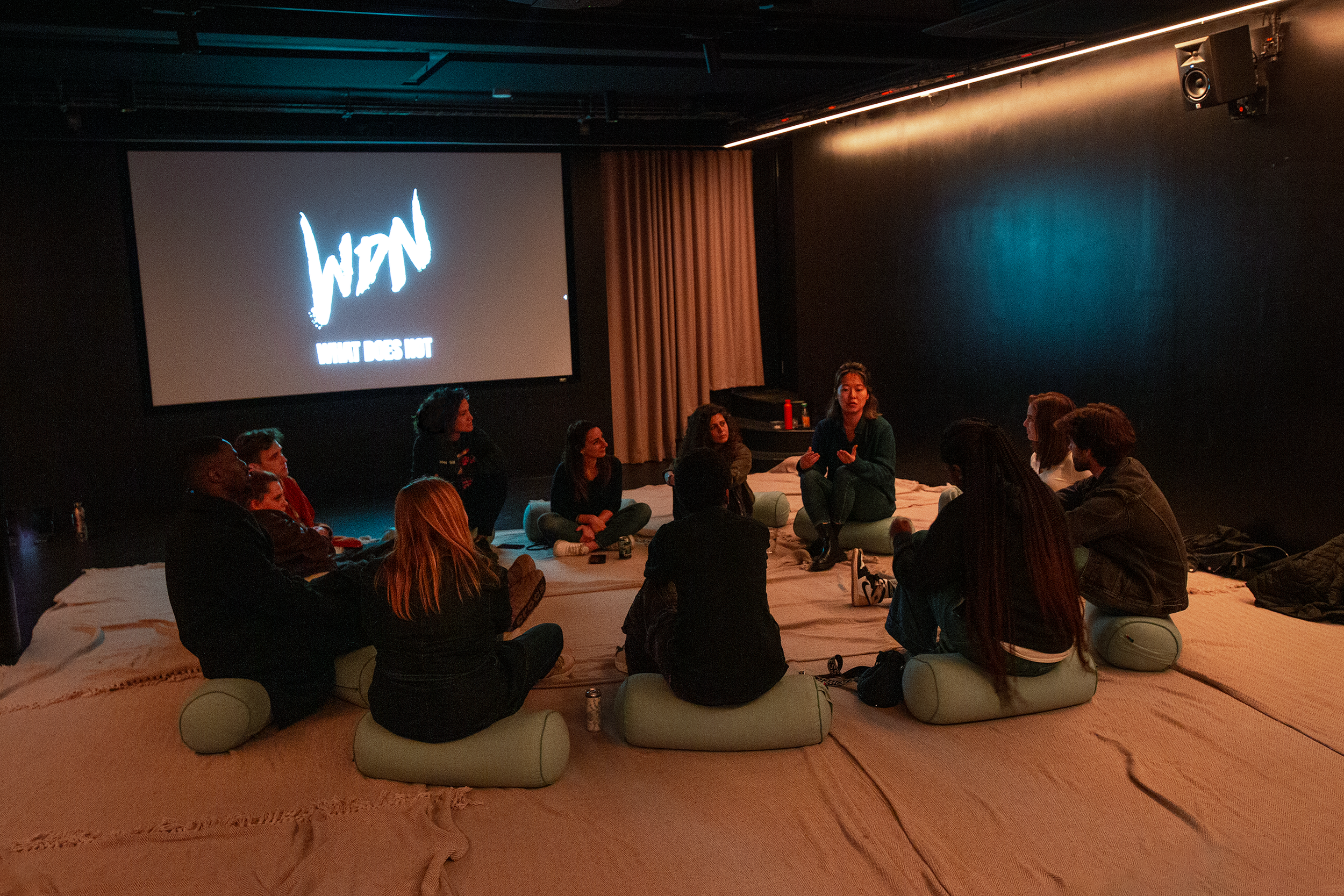 A group of people sitting on cushions in a dimly lit room, engaged in a discussion at a What Does Not WIP night, a Work in Progress night where musicians share projects before their release for audience feedback.