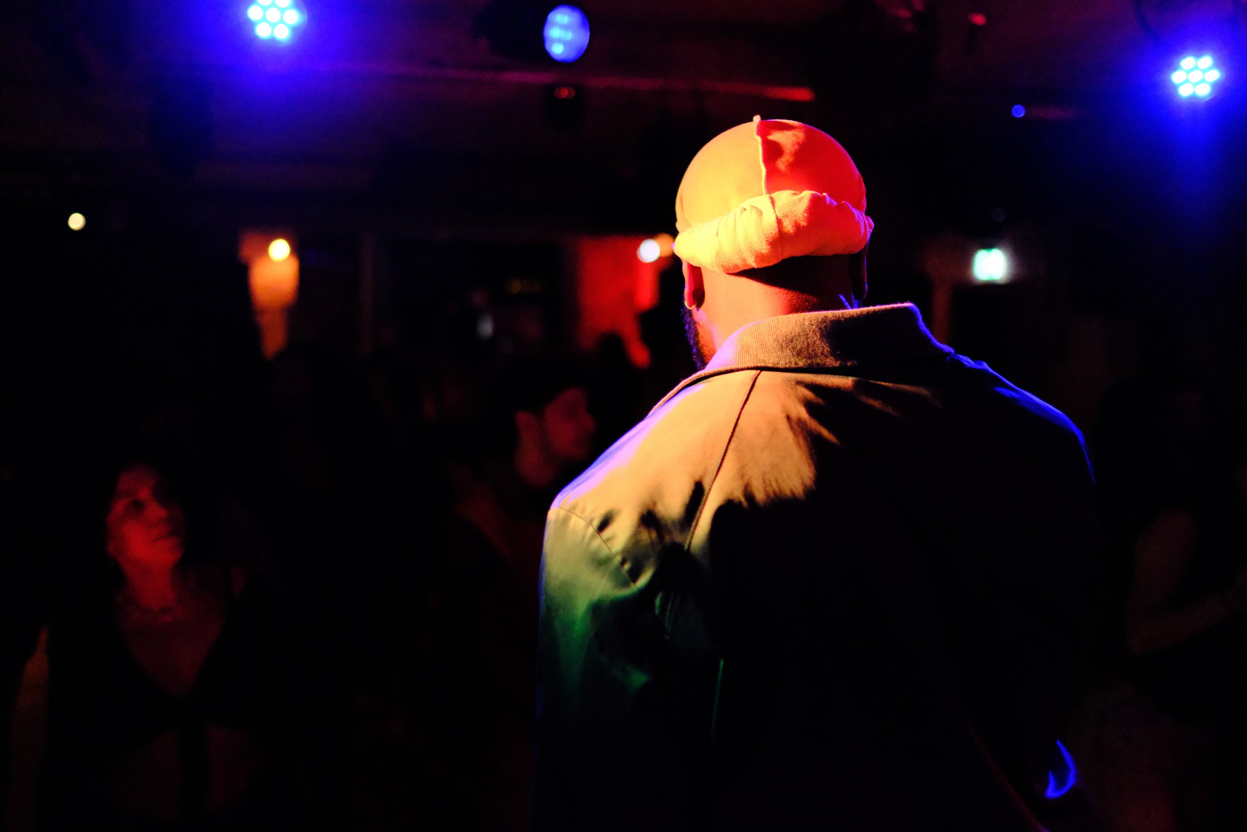 Emerging musician and rapper wearing a doorag, facing a dark room with colorful stage lights, surrounded by a crowd at a What Does Not emerging music showcase. 