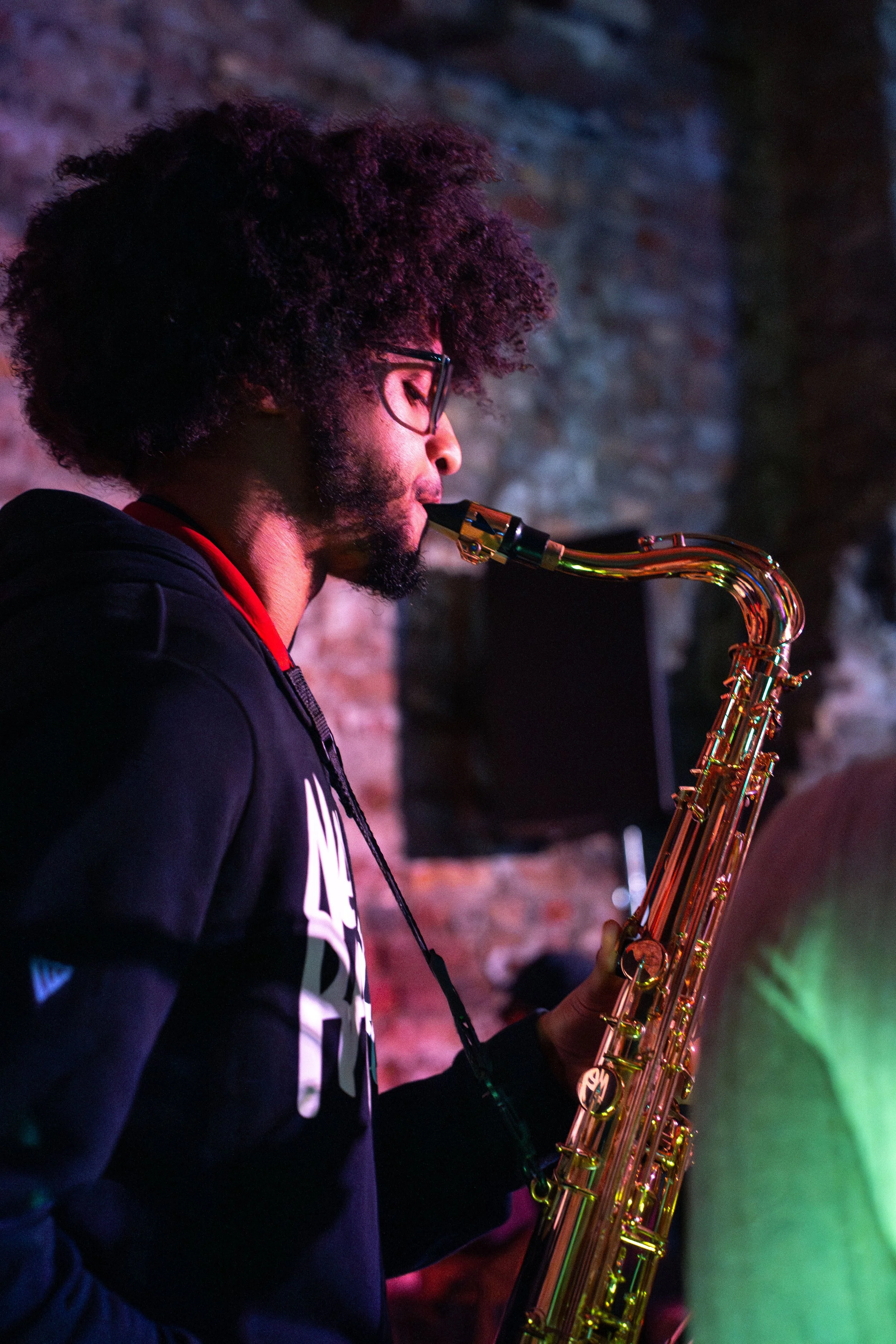 A man with glasses and curly hair playing a saxophone in a dimly lit setting with colorful lights and brick wall background.
