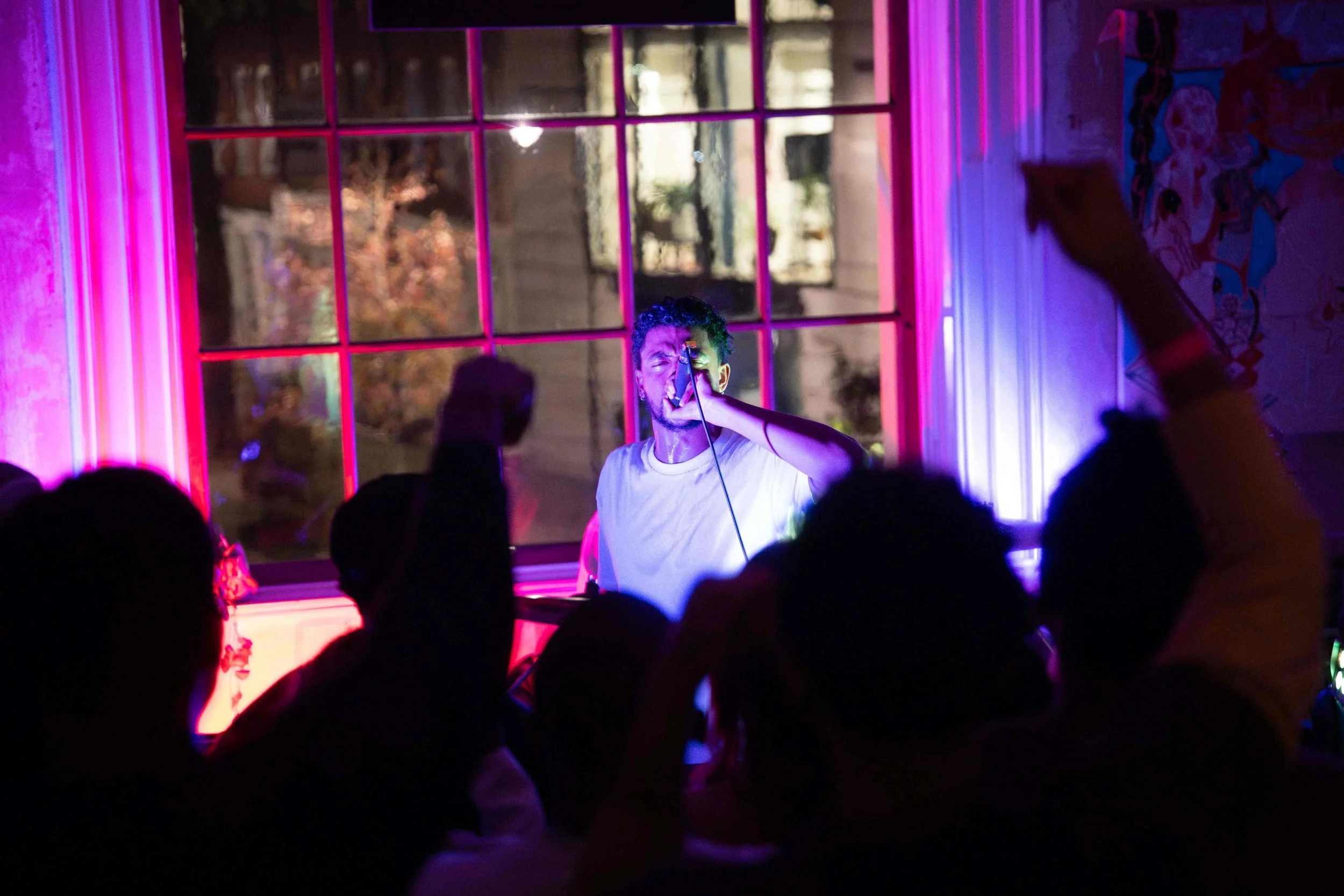 A man singing into a microphone during a live performance with a colorful, illuminated stage and audience in foreground at a RAW uncensored storytelling night in London