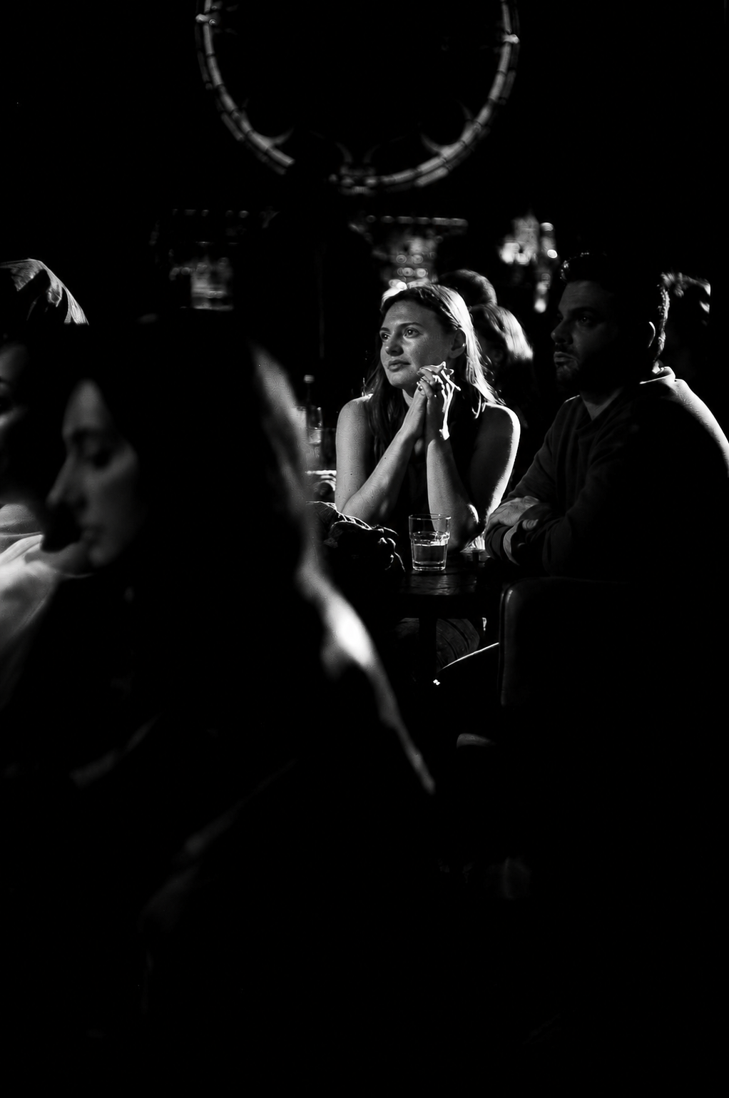 Black and white photo of people sitting at tables at a RAW uncensored storytelling night in a dark, dimly lit venue, with a woman in the center resting her chin on her hands, looking contemplative as she listens to true stories by everyday people