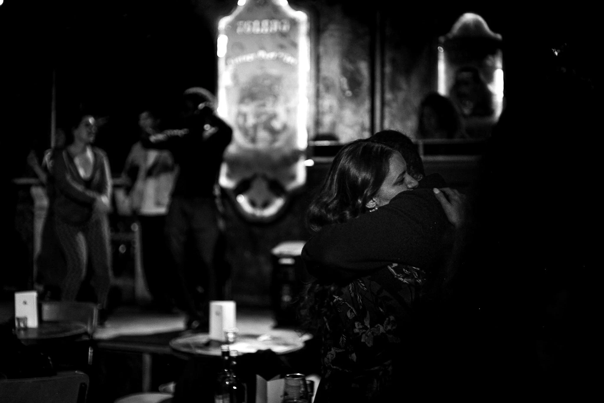 People hugging at a RAW uncensored storytelling night with tables and bottles in the foreground, and blurred figures in the background.