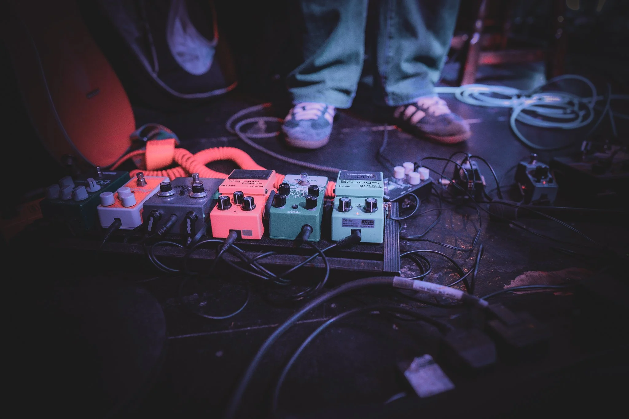 Close-up of guitar effects pedals connected with cables on a dark stage floor, with a person standing nearby wearing casual shoes and jeans.