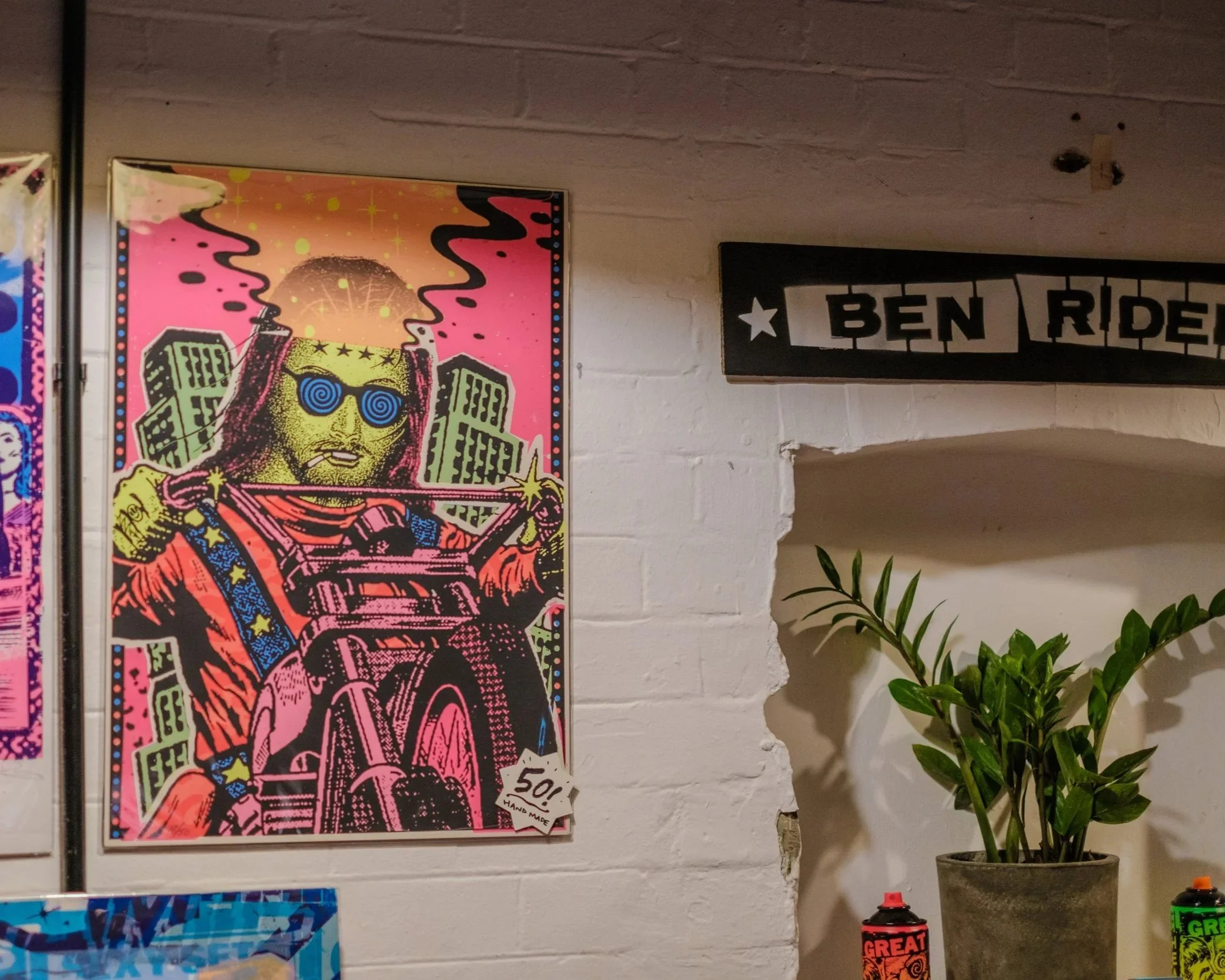 Art from the pop-up makers' market at a RAW uncensored storytelling night. Colorful pop art style poster of a person playing a guitar, wearing sunglasses, with buildings and starry effects in the background, hung on a white brick wall