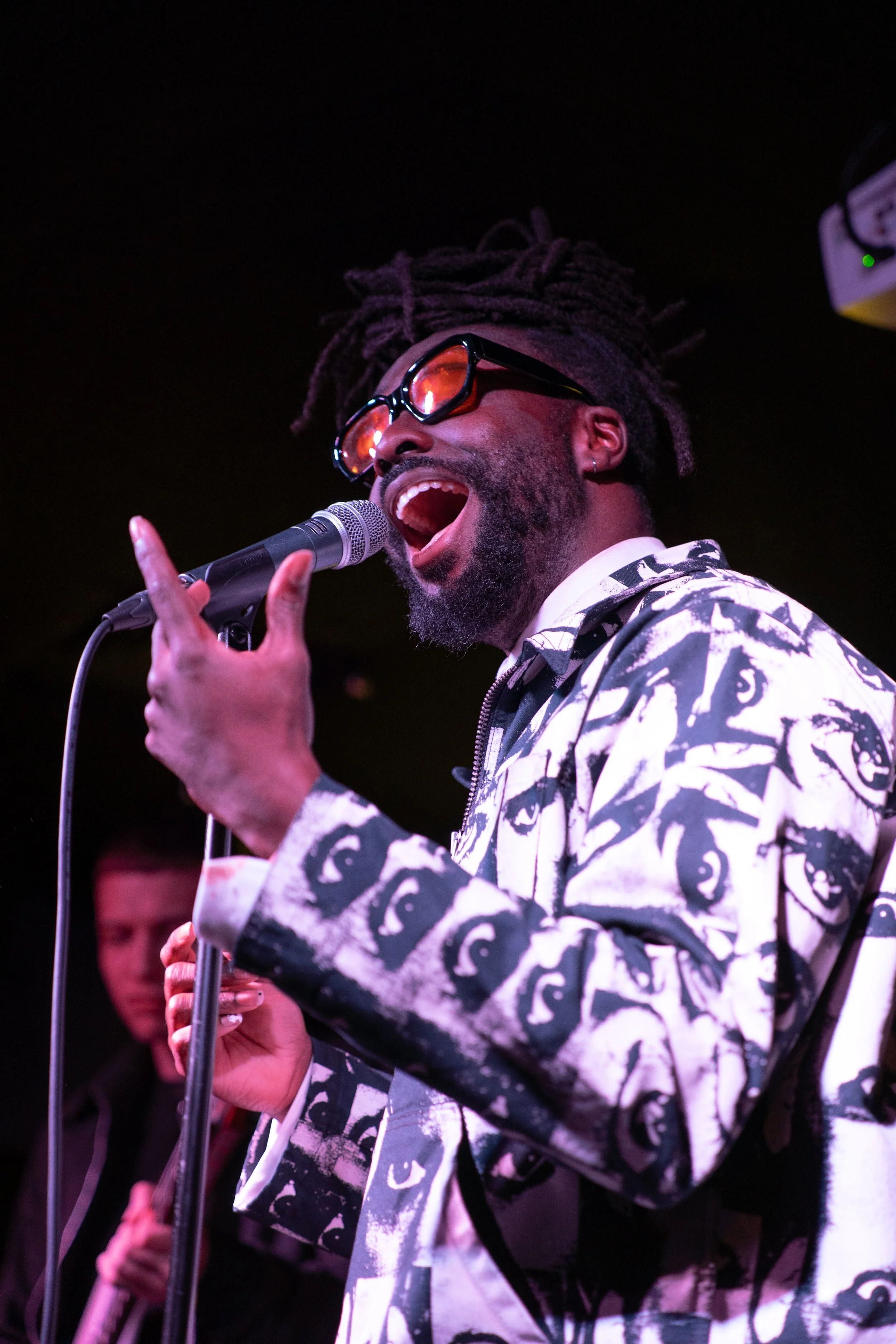 An emerging musician with dreadlocks, wearing glasses and a patterned jacket, sings into a microphone during a What Does Not emerging musician showcase.