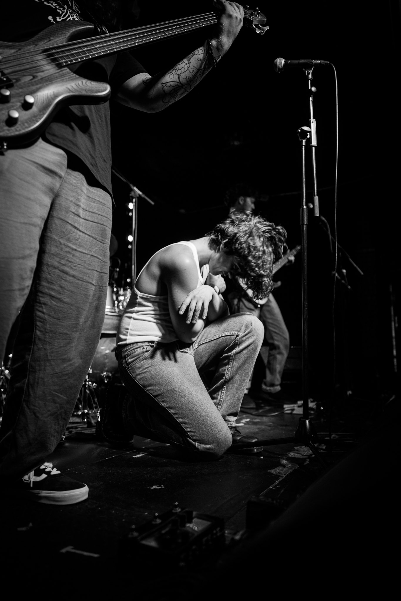 A black and white photo of a live concert in London by What Does Not, showing an independent musician and his band sitting on the floor with her head bowed, surrounded by musicians, one of whom is playing an electric bass guitar. 
