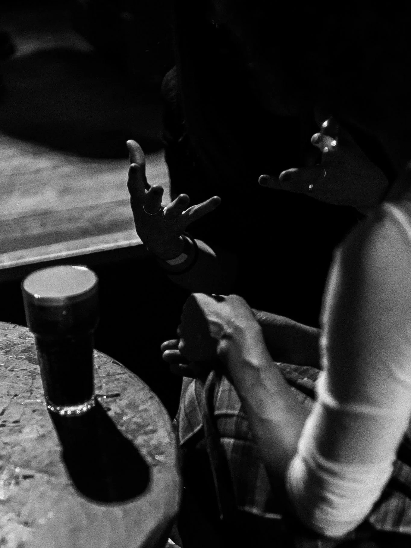 A moment between audience members captured at a RAW uncensored storytelling night in black and white photo of hands resting on a table with several glasses of dark-colored drinks.