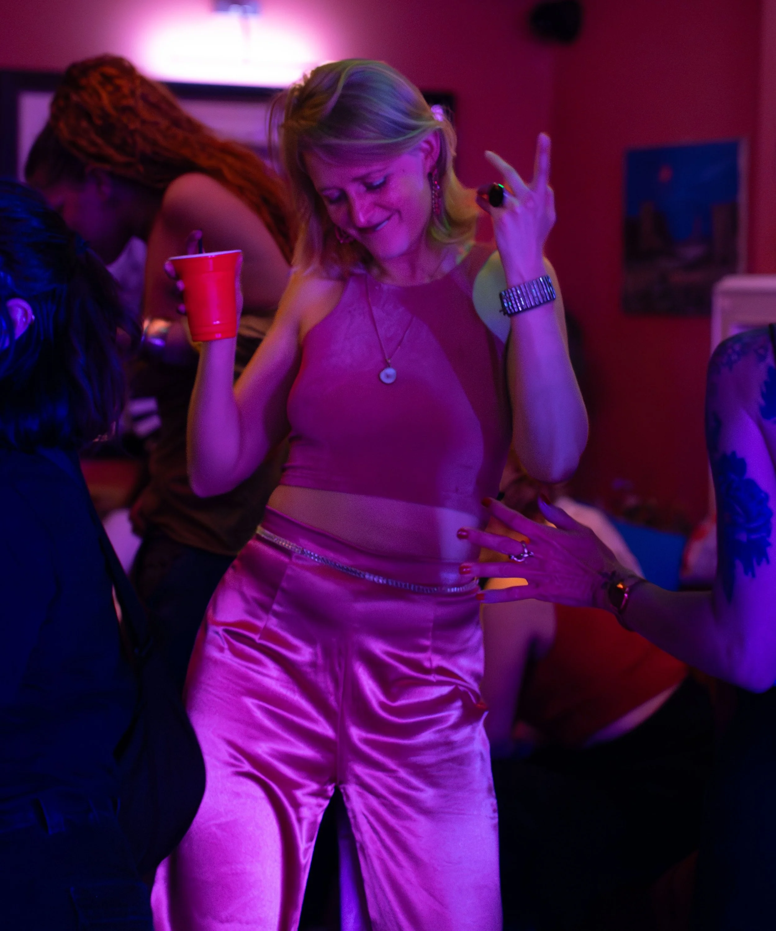 A woman dancing at What Does NOT house party with colorful lighting, wearing pink satin pants and a sleeveless top, holding a red cup, and making a gesture with her hand.