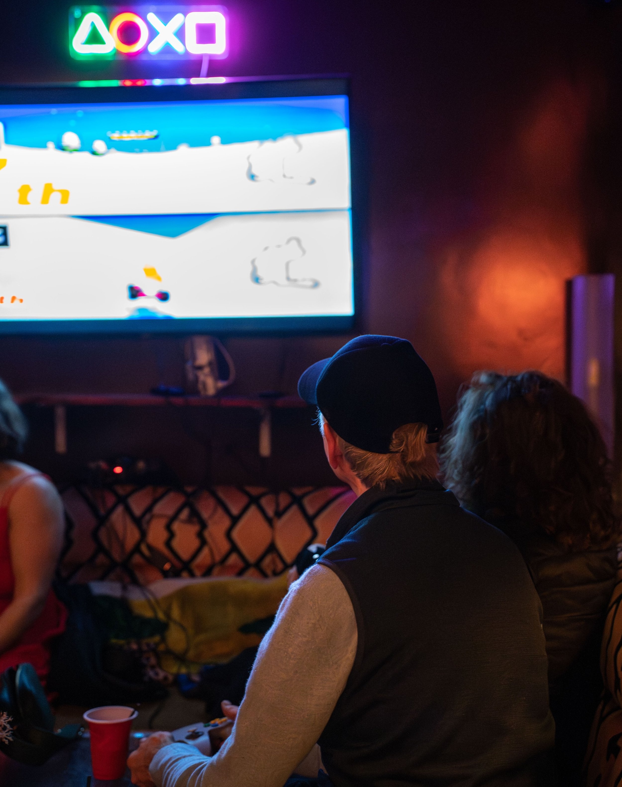 Group of people playing video games on television in a dark room with neon PlayStation symbols on the wall at a What Does Not house party in London