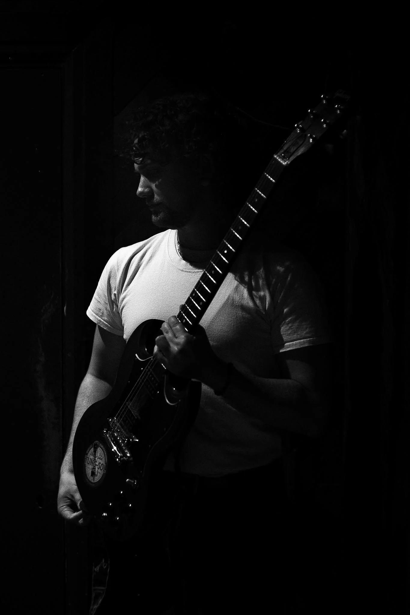 An emerging artist who performed at RAW uncensored storytelling in a white t-shirt playing an electric guitar in a dark room, with only outlines and the guitar's reflective parts visible.
