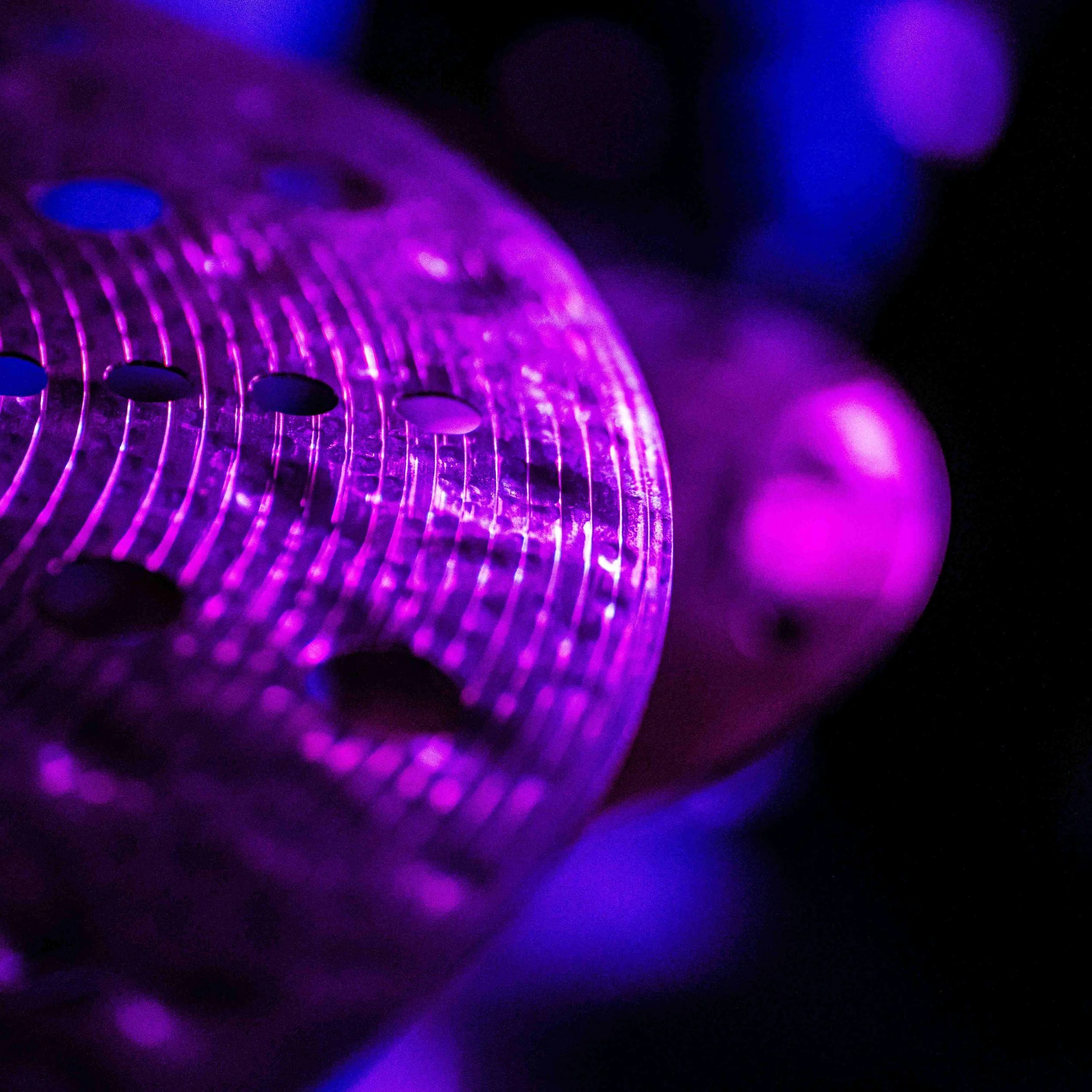 A close-up of a cymbal from a What Does Not music event, illuminated by purple and blue lights, with visible grille lines and circular holes.