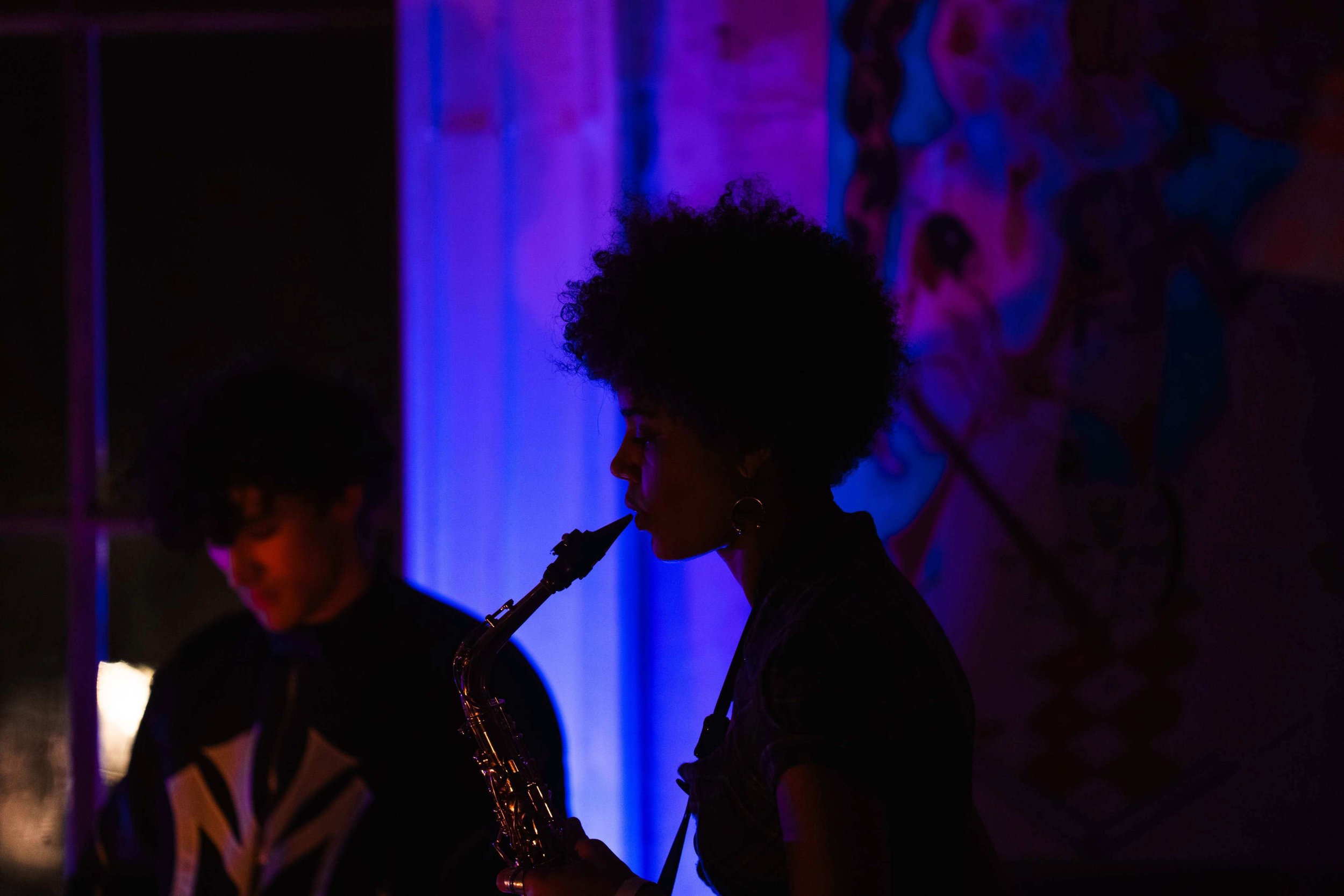 A person playing a saxophone in a dimly lit room with purple and blue lighting, and a colorful piece of artwork on the wall in the background at a What Does Not headline show for an emerging musician in London in support of grassroots music.